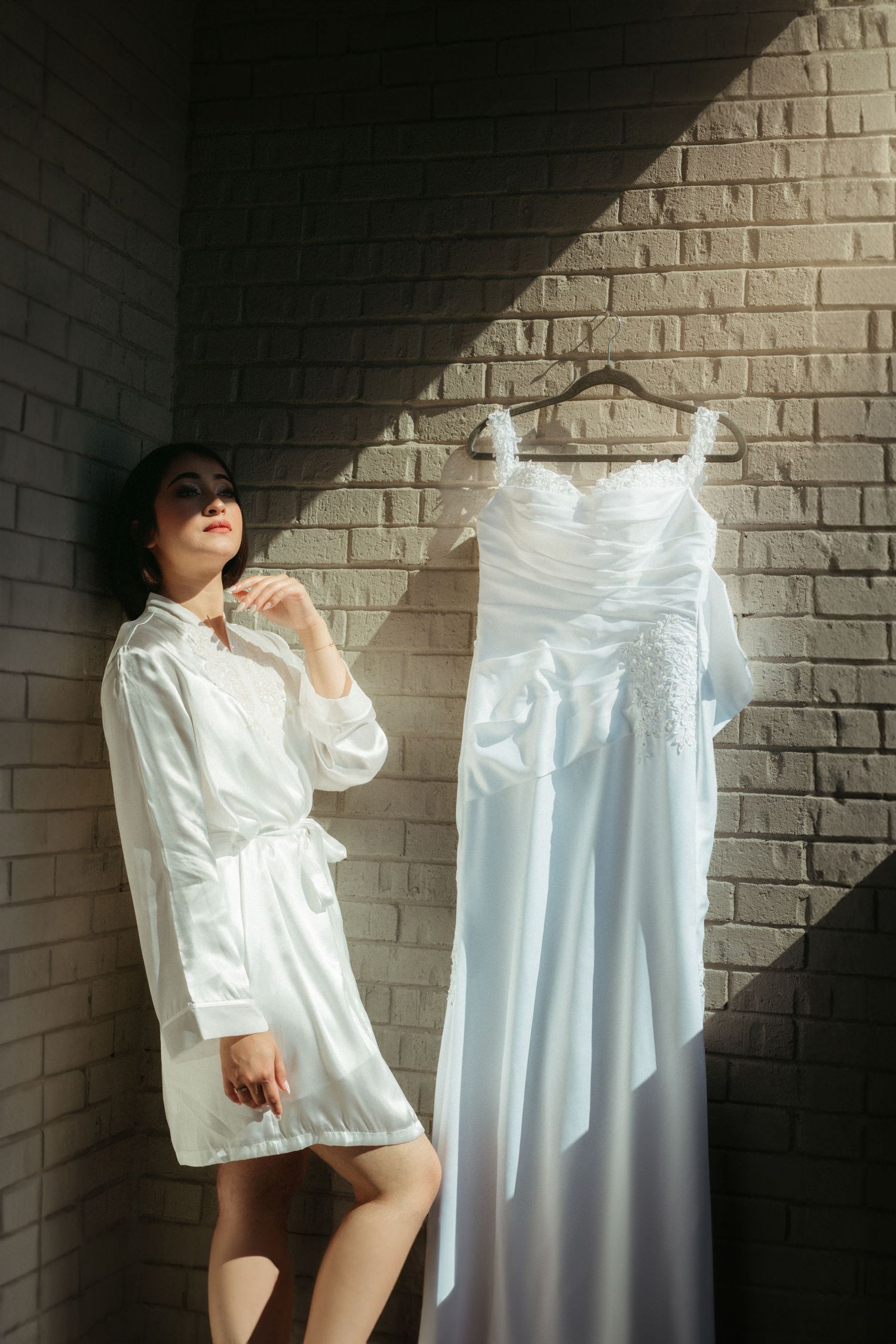 A white wedding dress hangs on the wall next to someone wearing a silky white robe in front of a brick background.