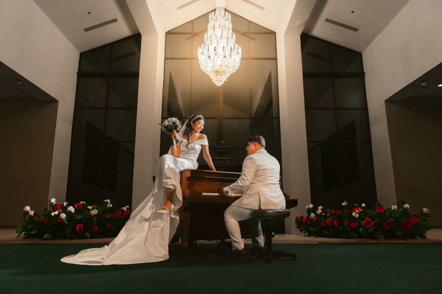 A wedding couple sits at a grand piano under an elegant crystal chandelier in a dramatic indoor setting.