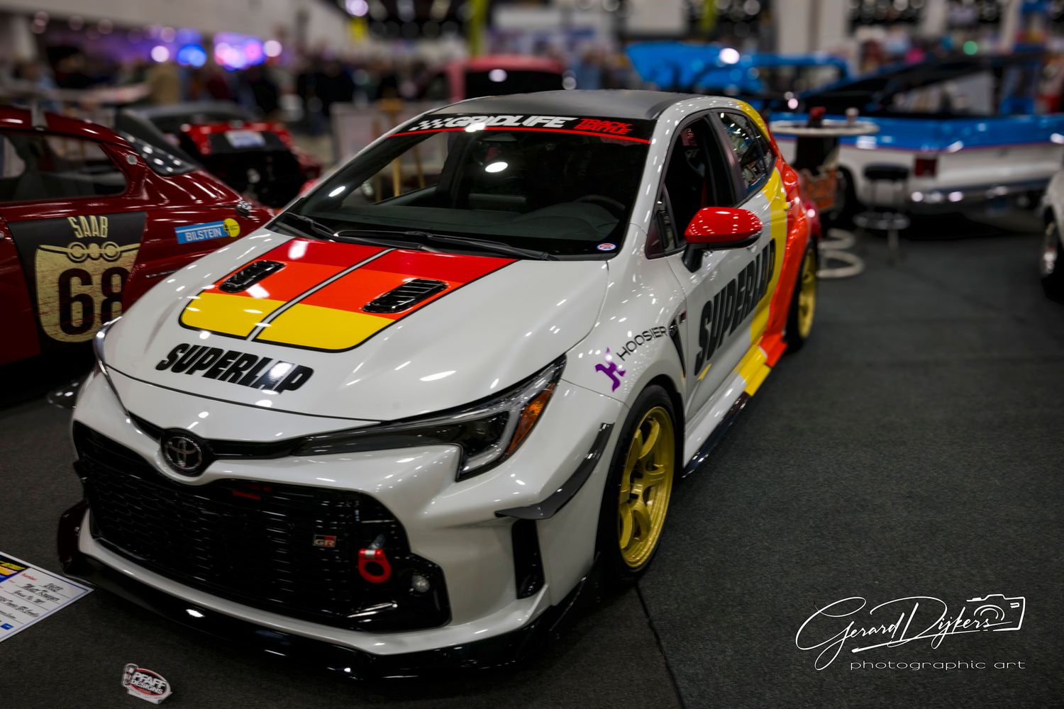 Modified Toyota GR Corolla race car with German flag livery and gold wheels at an indoor exhibition.