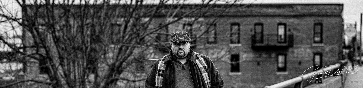 A person stands in front of a brick building in a black and white photograph wearing a winter coat.