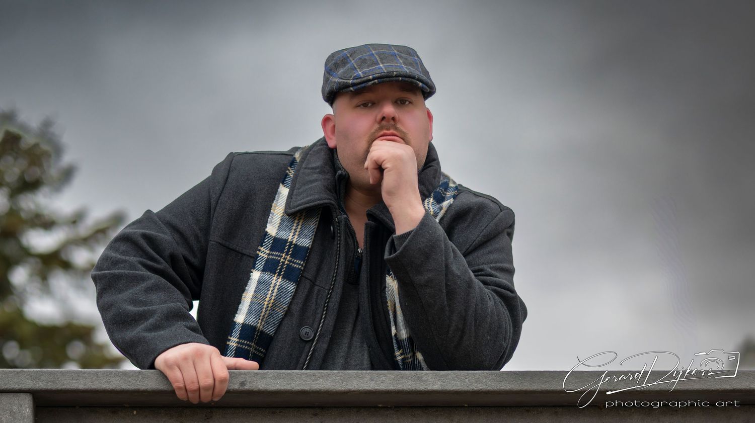 A person wearing a dark jacket and cap stands outdoors on a cloudy day looking through binoculars.