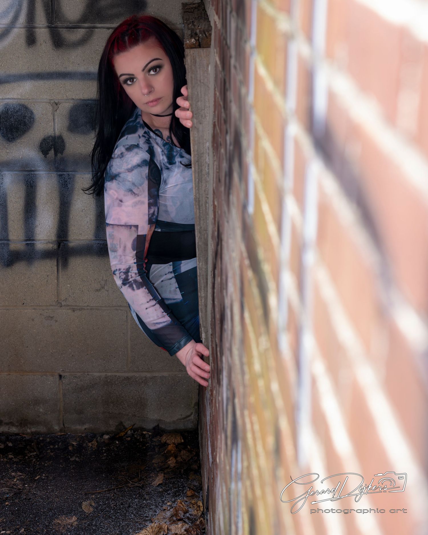 A person in a black jacket poses against a concrete wall with dramatic shadows and light creating an artistic effect.