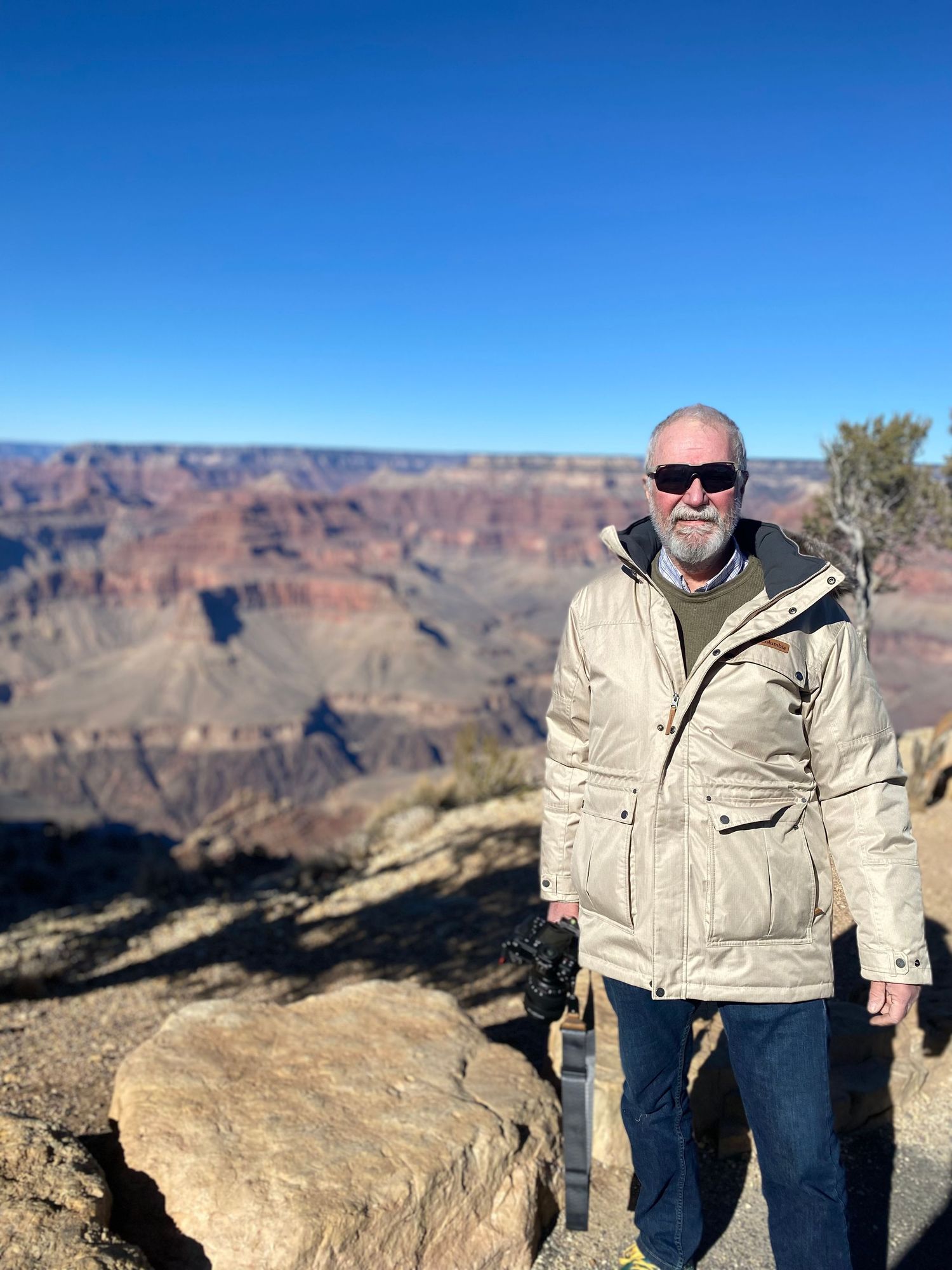 A person in a beige jacket and sunglasses stands at a Grand Canyon viewpoint overlooking the vast desert canyon landscape.