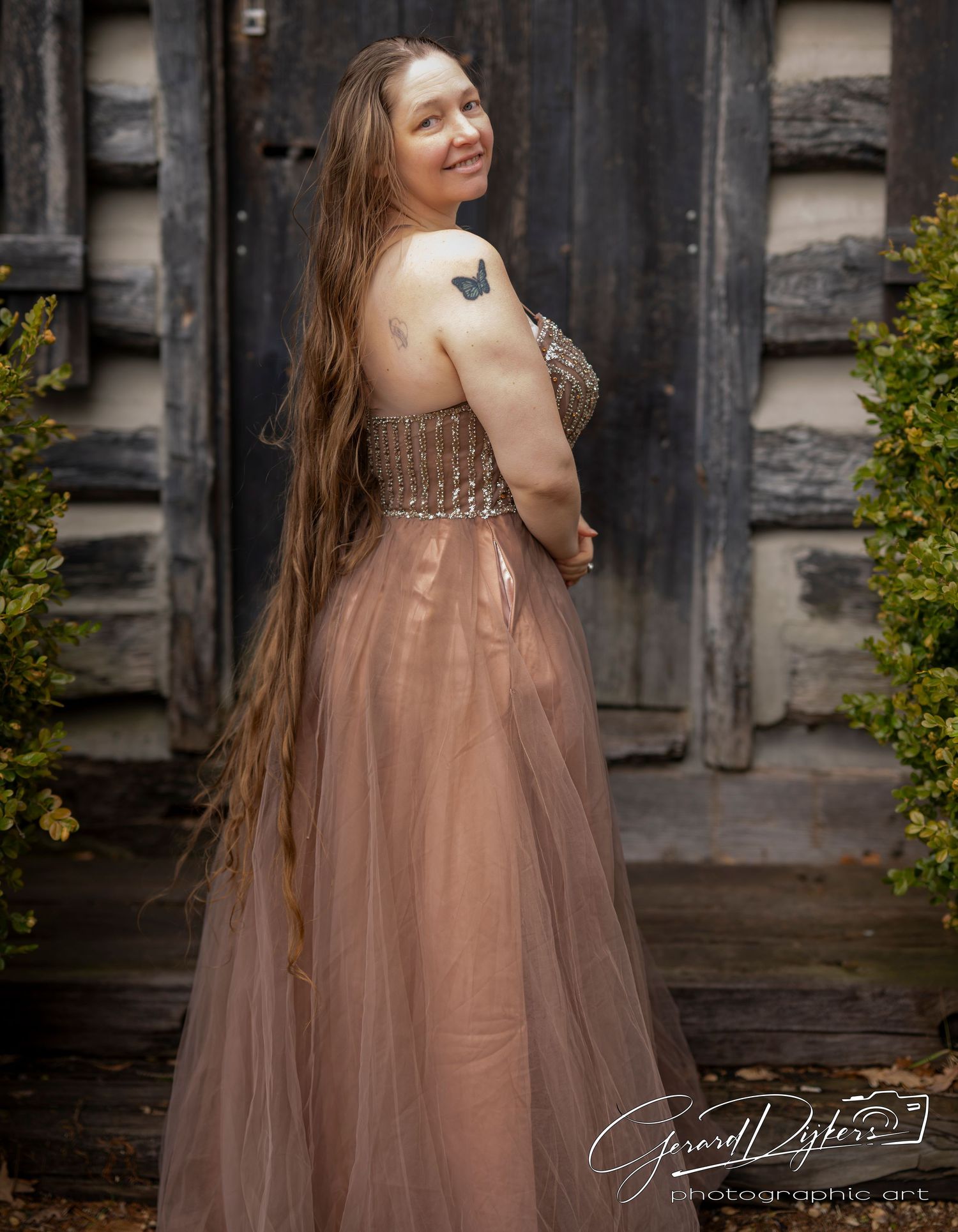 A woman in a flowing dusty rose tulle gown poses elegantly against a rustic dark wooden backdrop.
