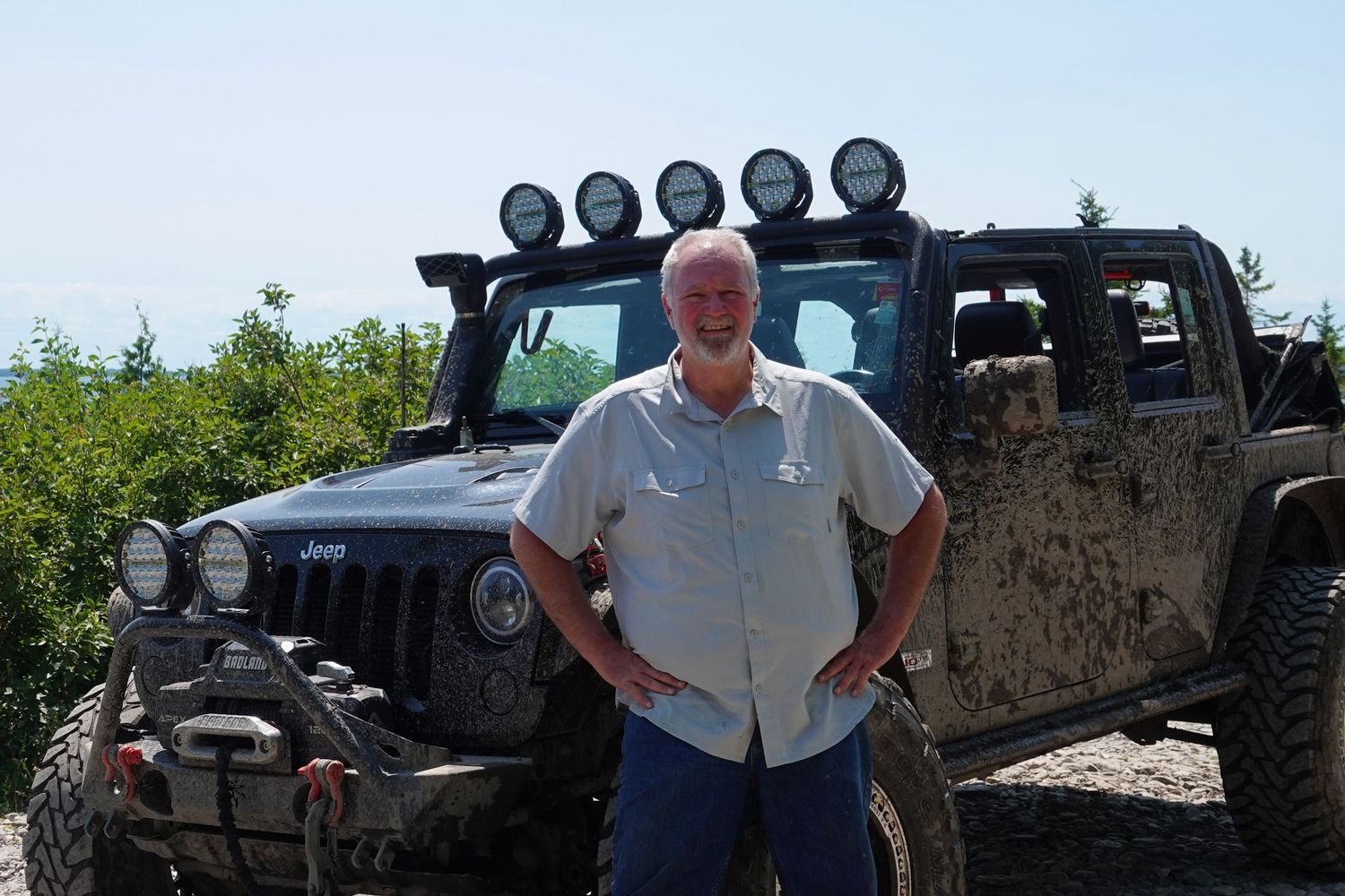 A muddy modified Jeep Wrangler with off-road lights, winch and lift kit parked outdoors in bright sunlight.