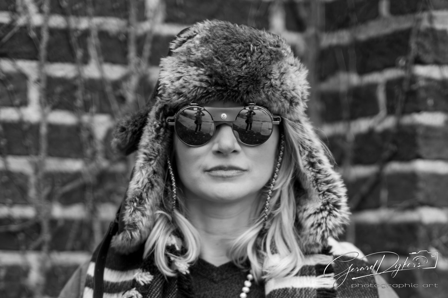 Person wearing winter fur hat and sunglasses against brick wall background.