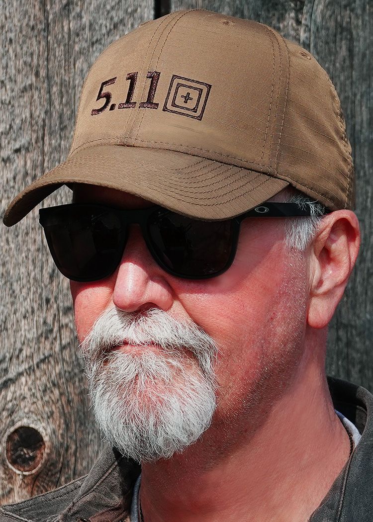 Close-up view of someone wearing a tan 5.11 baseball cap and dark sunglasses against a rustic wooden background.