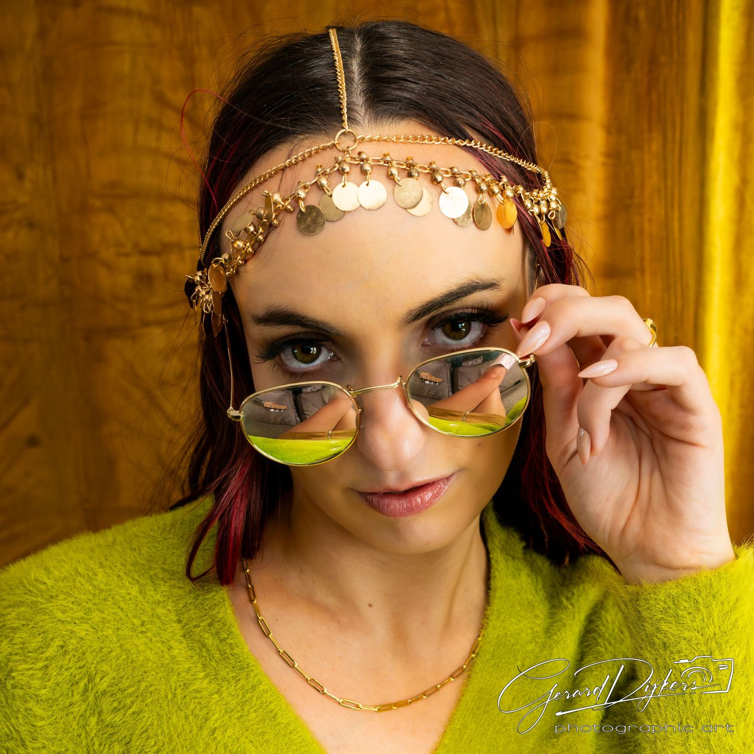Woman in bright lime green sweater wearing decorative gold headpiece and yellow-tinted sunglasses against golden background.