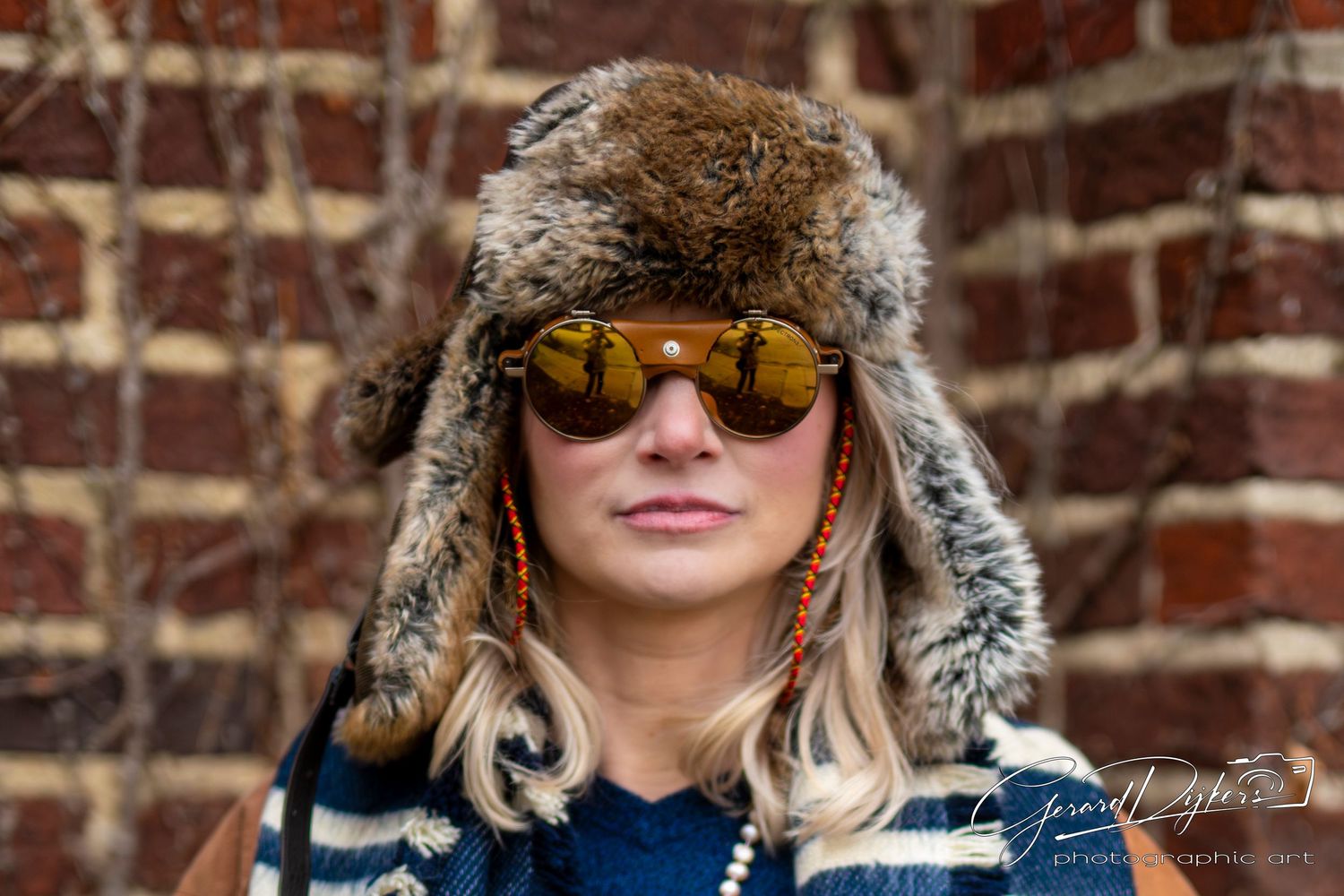 Close-up portrait of someone in fur trapper hat and aviator sunglasses with brick wall behind.