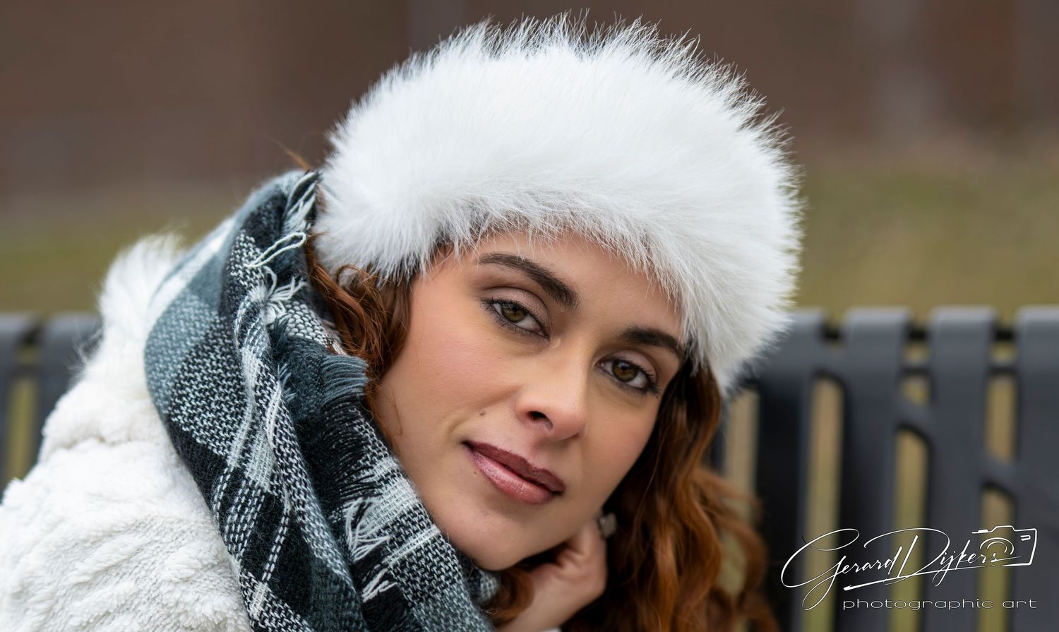 A person wearing a white furry winter hat and gray scarf poses in an outdoor setting during winter.