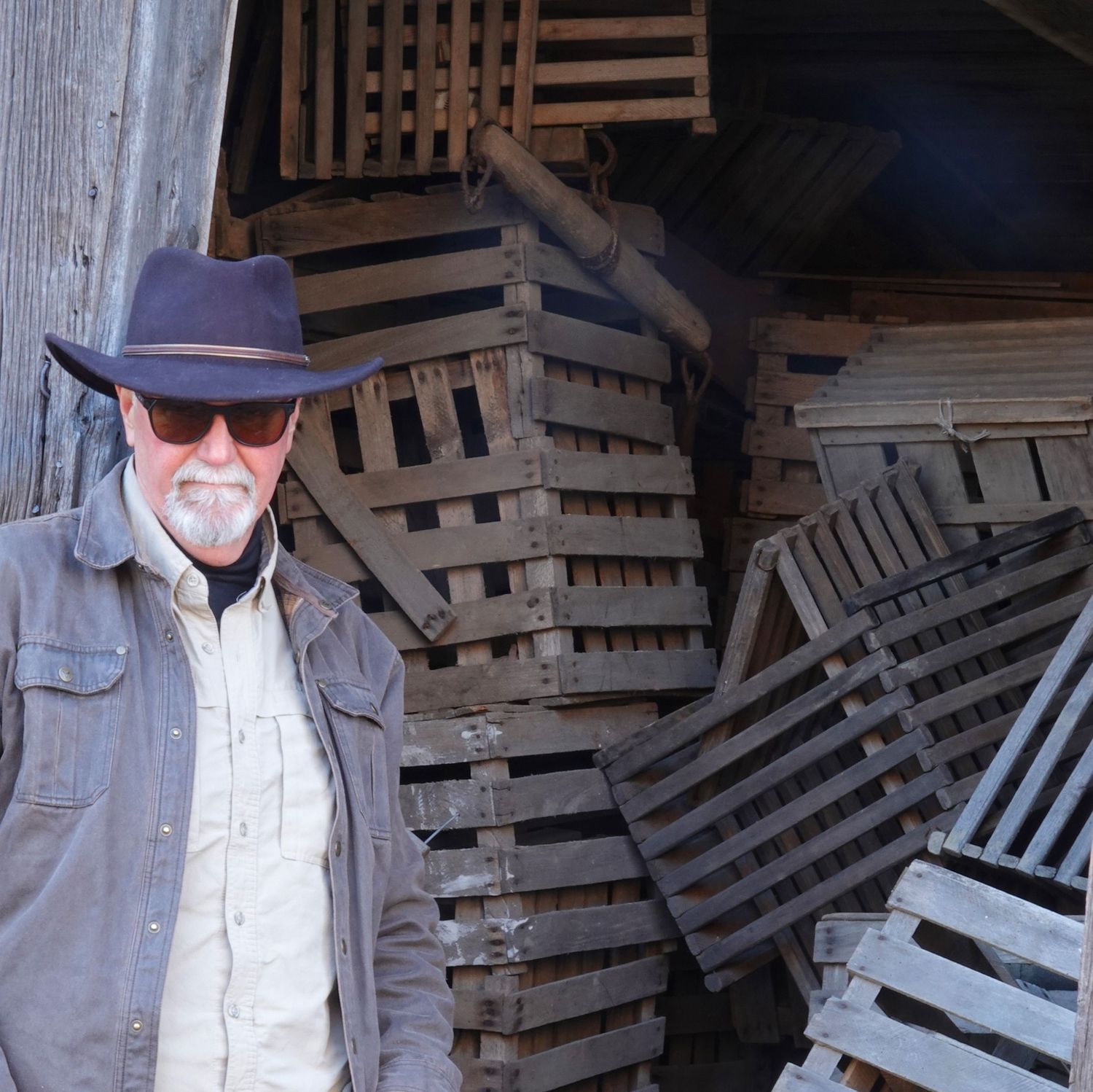 Person wearing brown jacket and hat standing in front of stacked wooden pallets in dimly lit storage area.