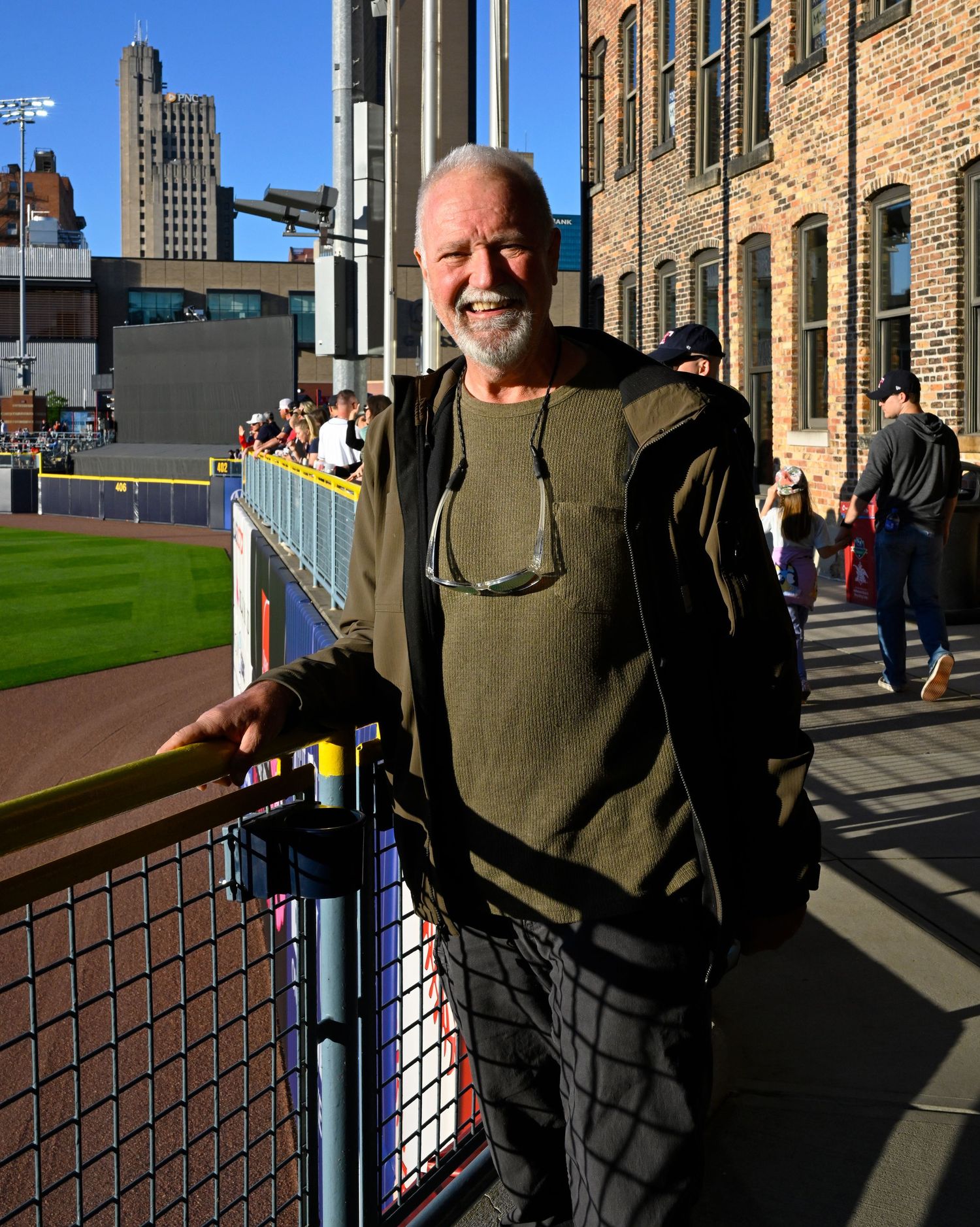 A person in olive green sweater stands smiling along a baseball stadium railing on a sunny day.