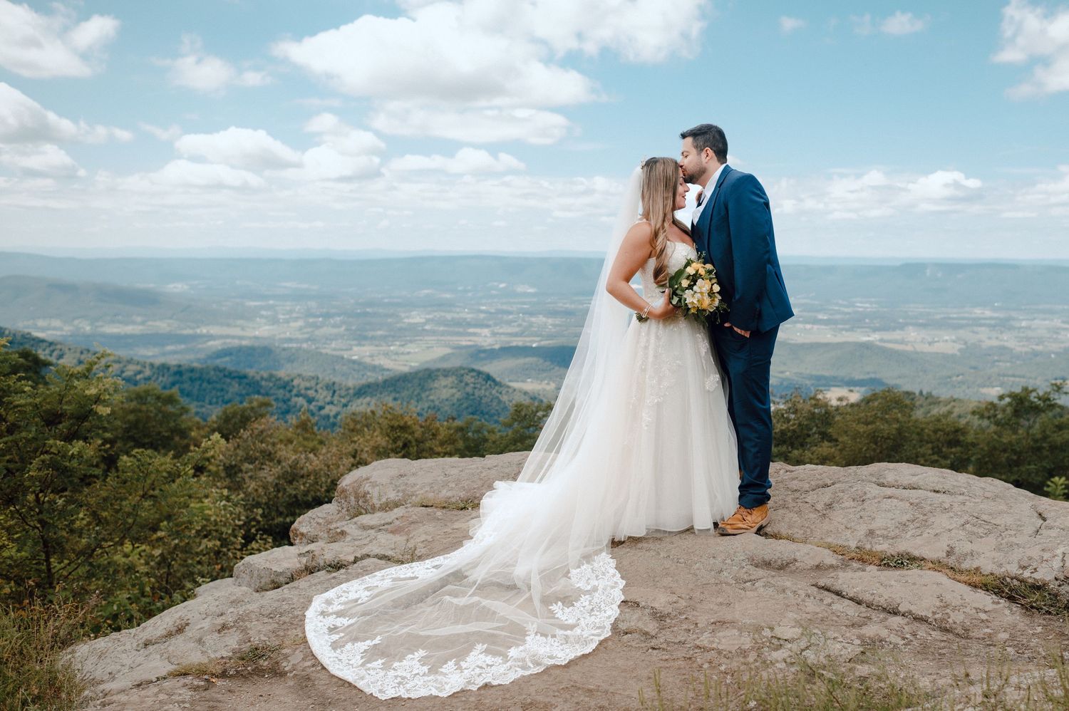 Two people in wedding attire stand together on a rocky mountain overlook with a scenic valley vista stretching below.