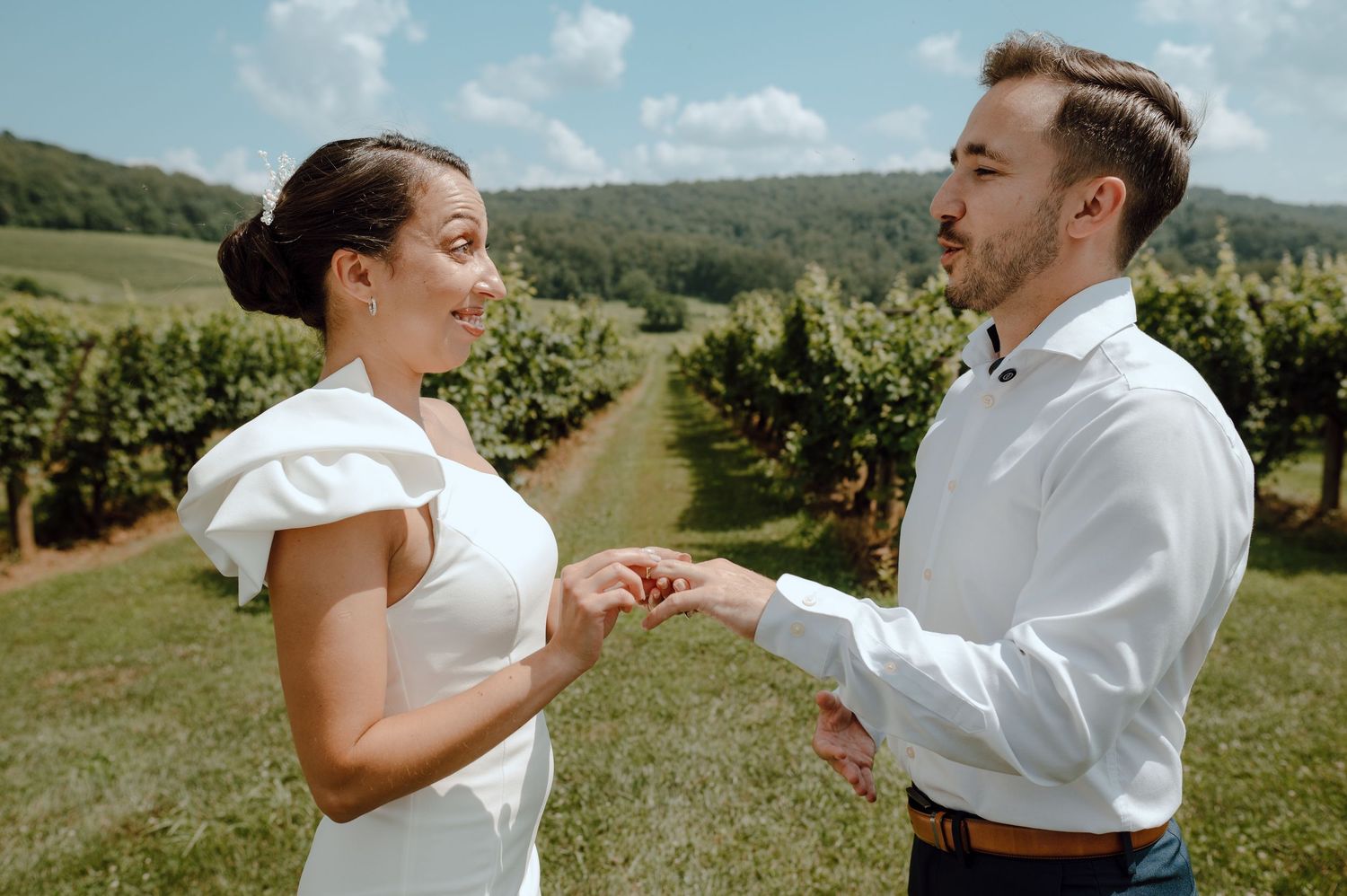 A couple in wedding attire hold hands while standing in a scenic vineyard with rolling hills and trees in the background.