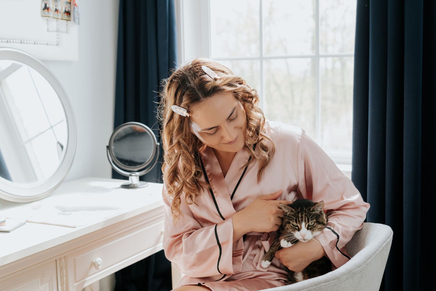 Person in pink pajamas sits by a window holding a small kitten in a bright, airy room.