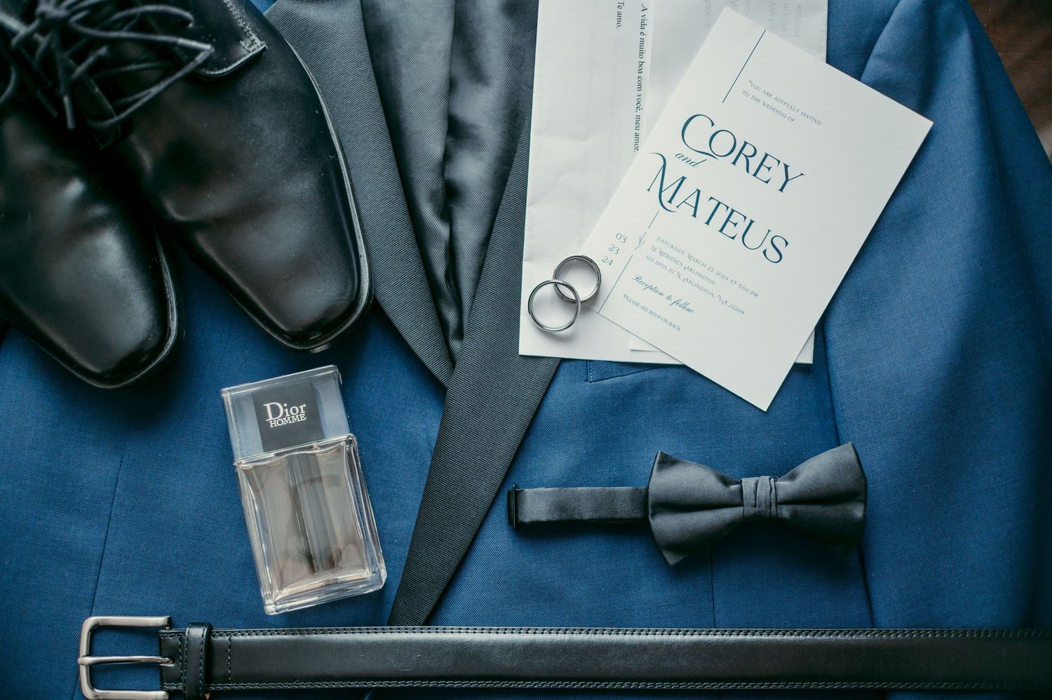 Wedding accessories including blue suit, black shoes, bow tie, belt and nuptials document laid out flat.
