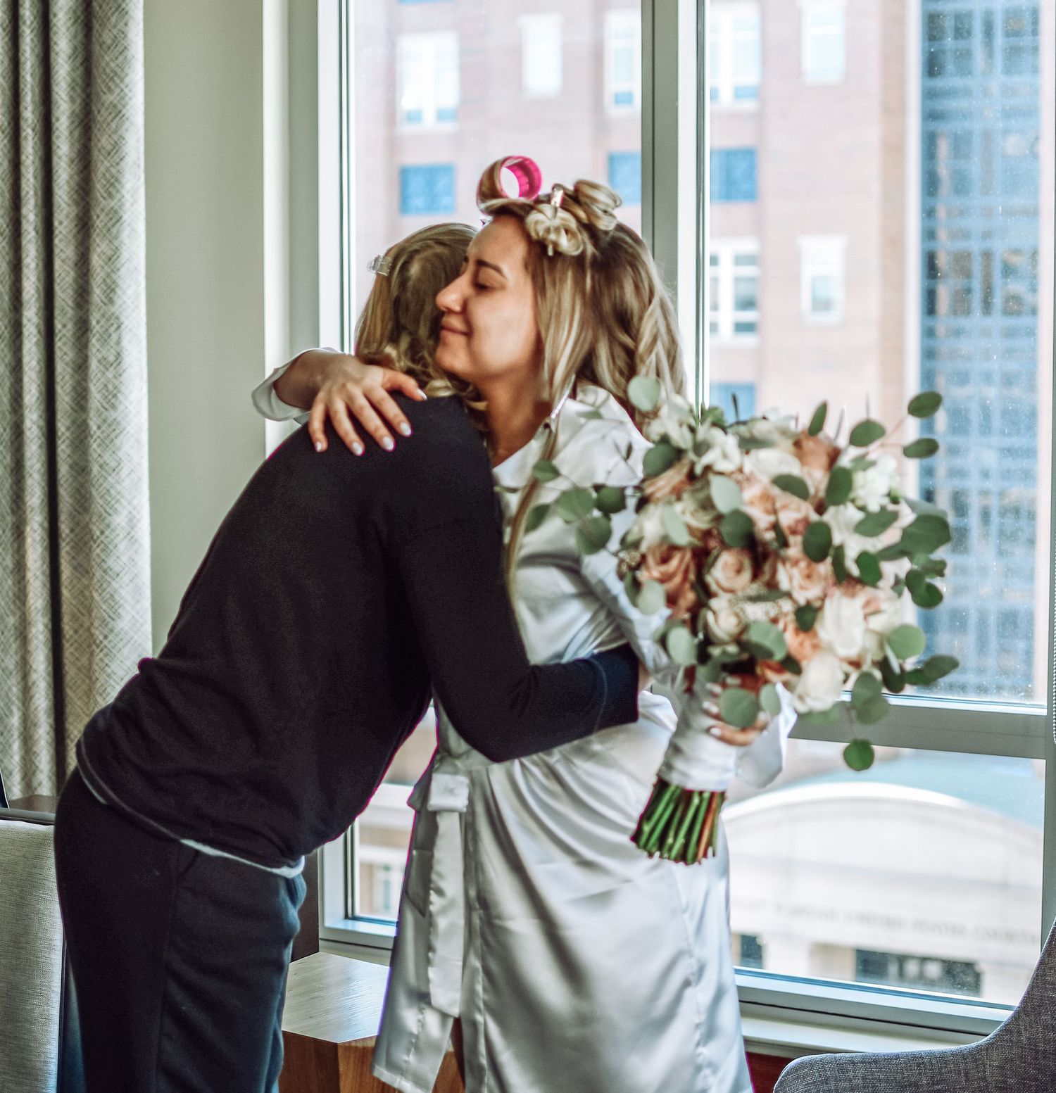 Two people embrace in a tender moment near bright windows with a floral bouquet in an indoor setting.