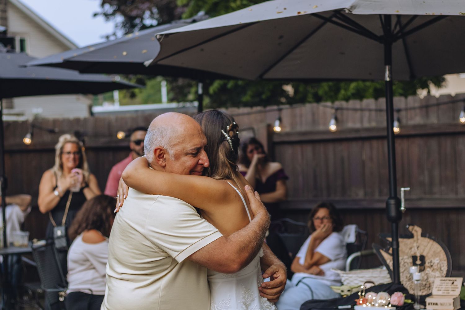 Two people embrace in an emotional hug during an outdoor gathering with patio umbrellas and guests looking on.