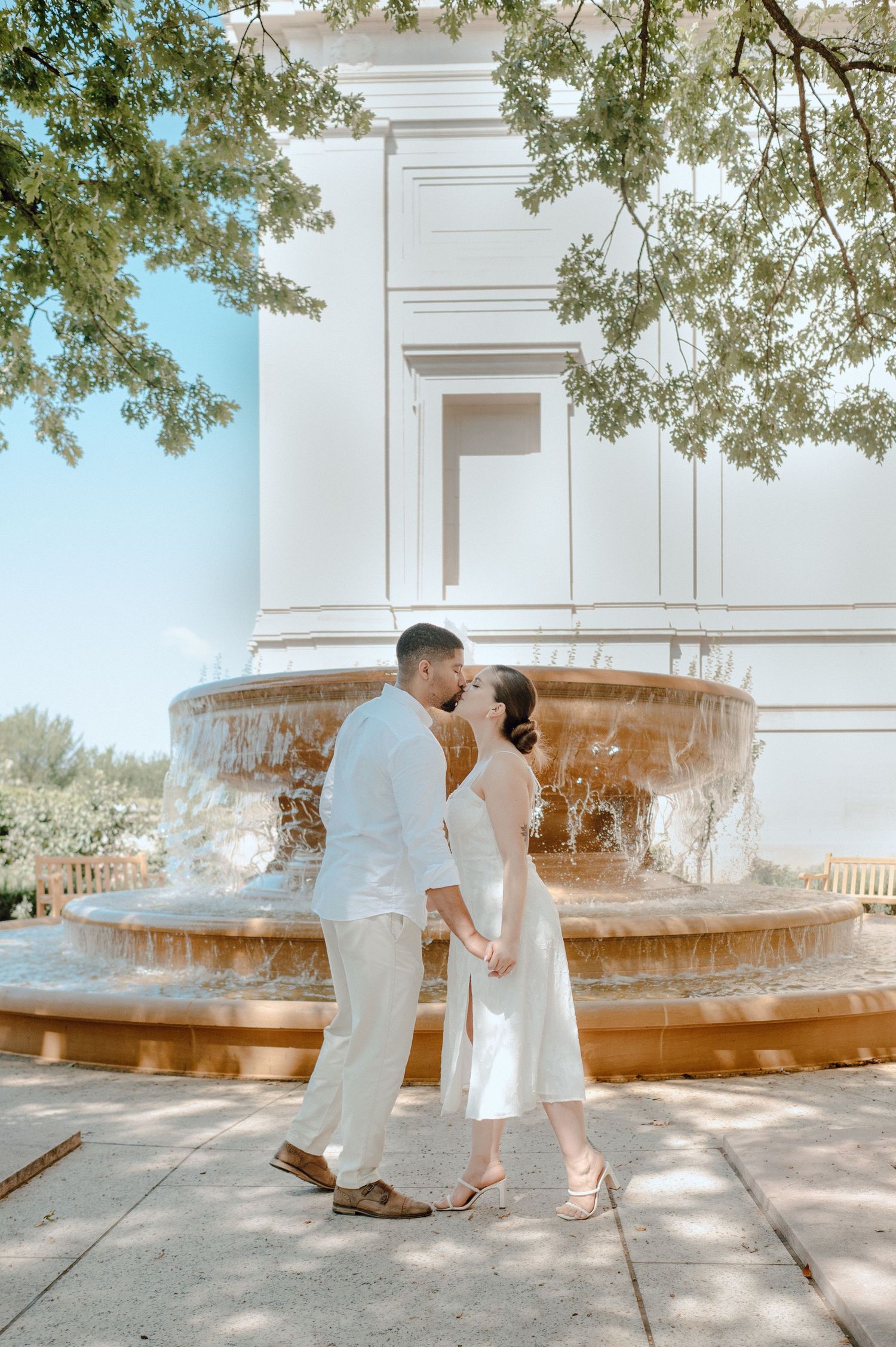 A romantic photoshoot by a fountain showing tender moments between a couple in white outfits on a sunny day.