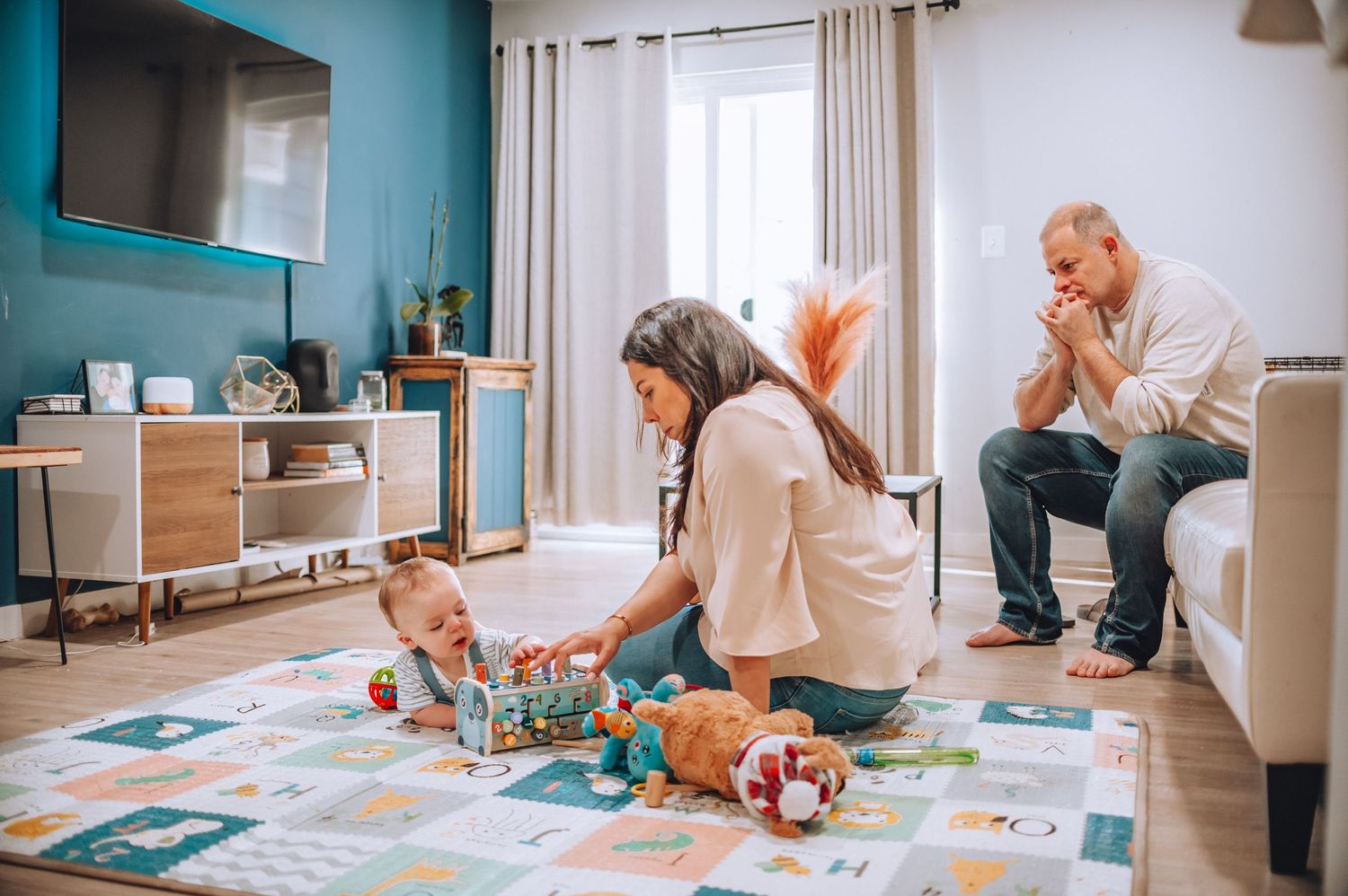 Family gathered in living room with turquoise wall playing with baby on floor mat.