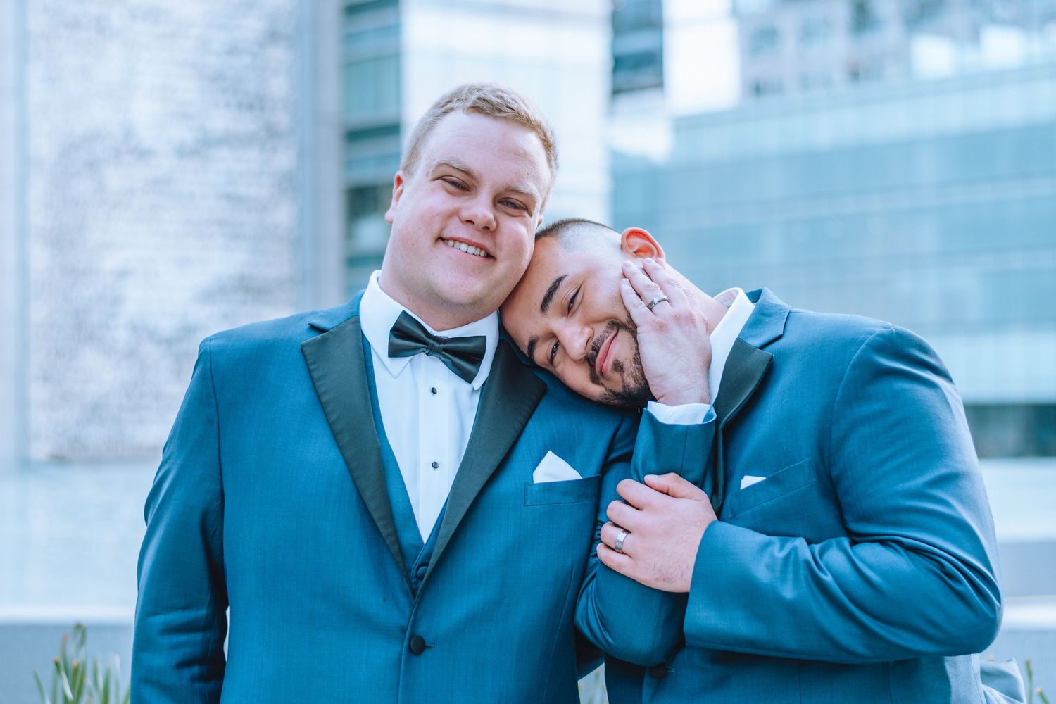 Two people wearing teal blue suits and bow ties embrace while posing together for a formal wedding photo.