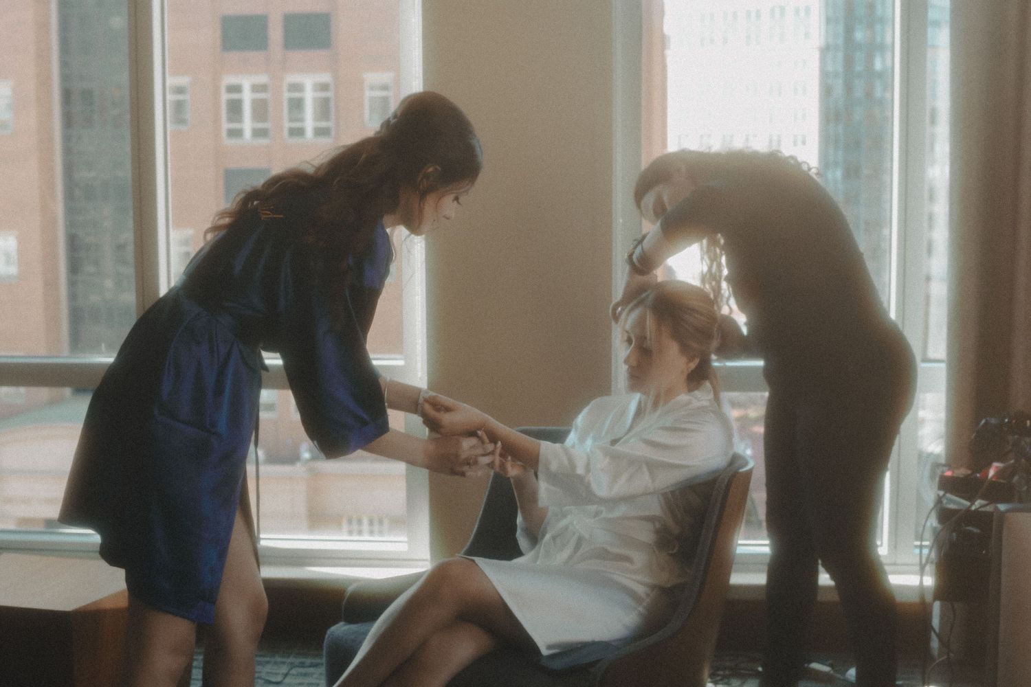 Three people in a sunlit room during a hair and makeup styling session near tall windows in an urban setting.