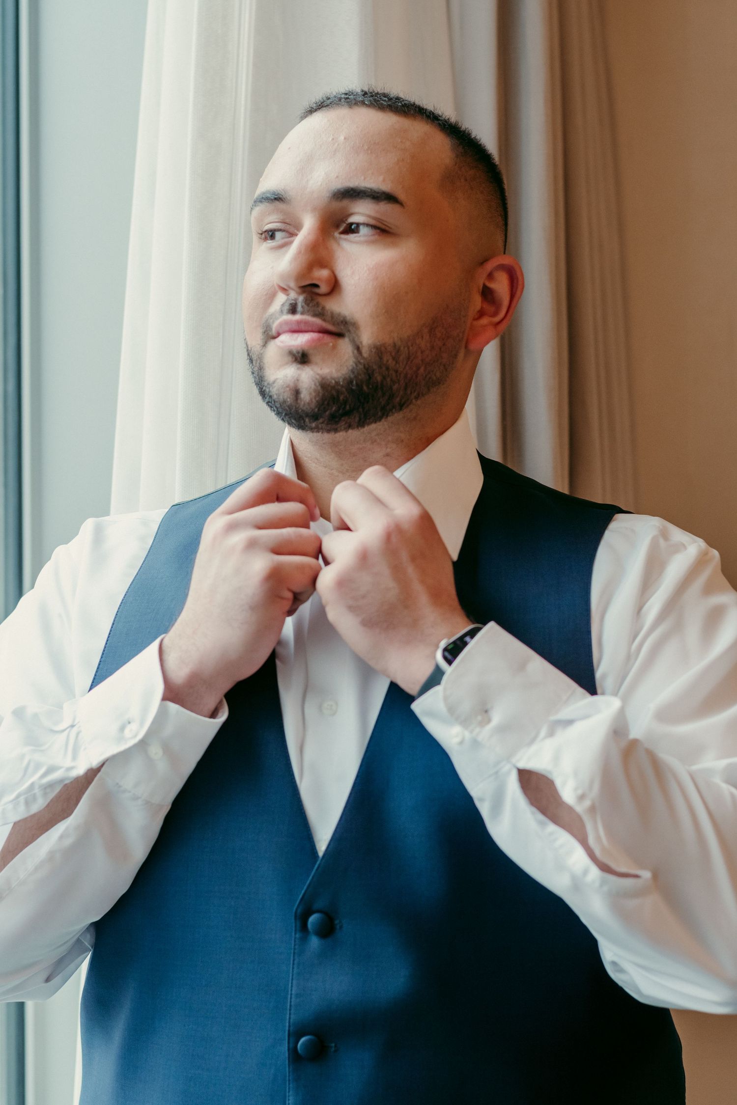 A groom in a navy blue vest and white shirt adjusts his tie while standing by a window in natural light.