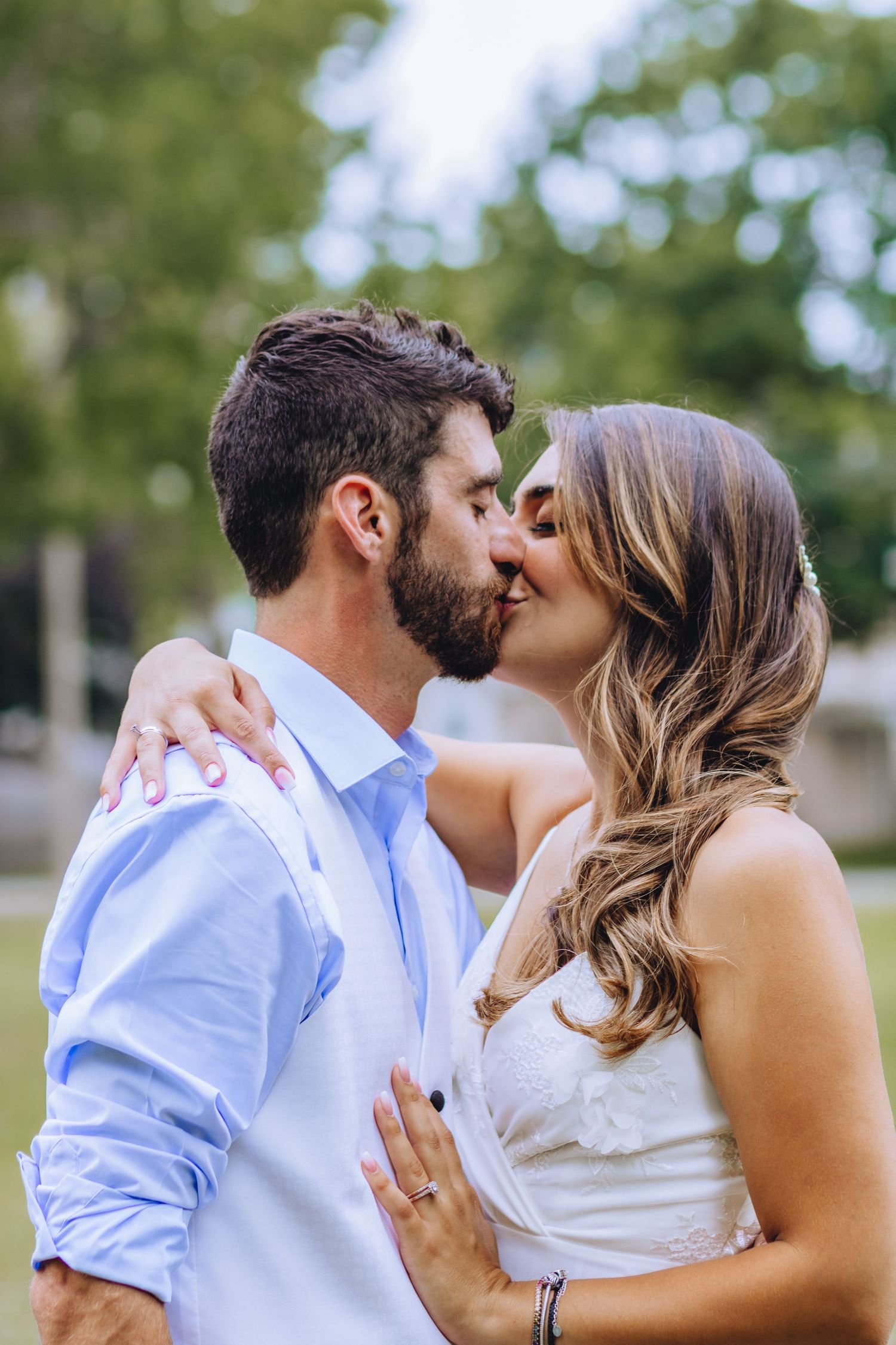 A couple shares a romantic kiss outdoors with soft bokeh and greenery in the background.