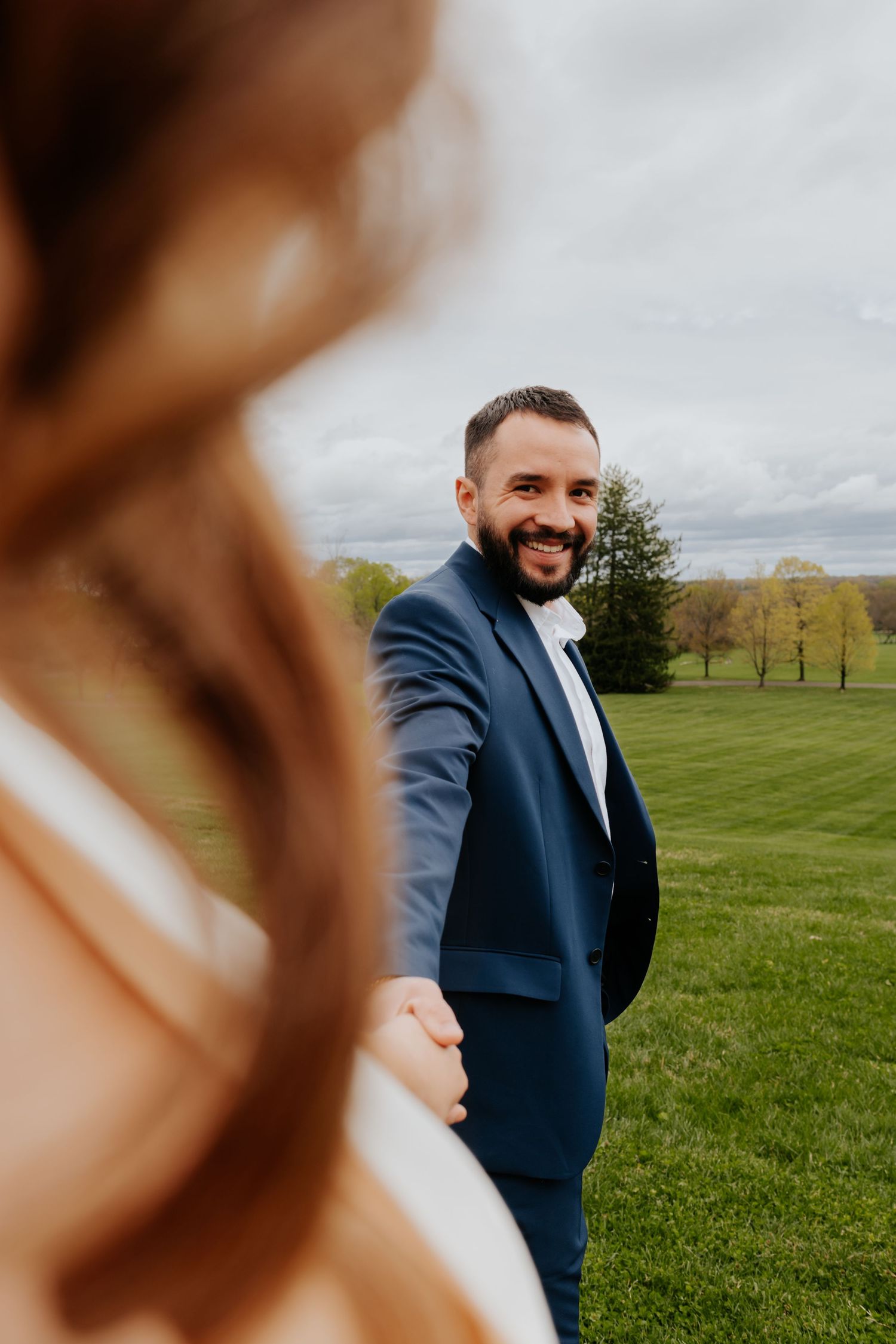 A joyful couple holds hands in a grassy field with cloudy skies behind them during what appears to be their wedding day.