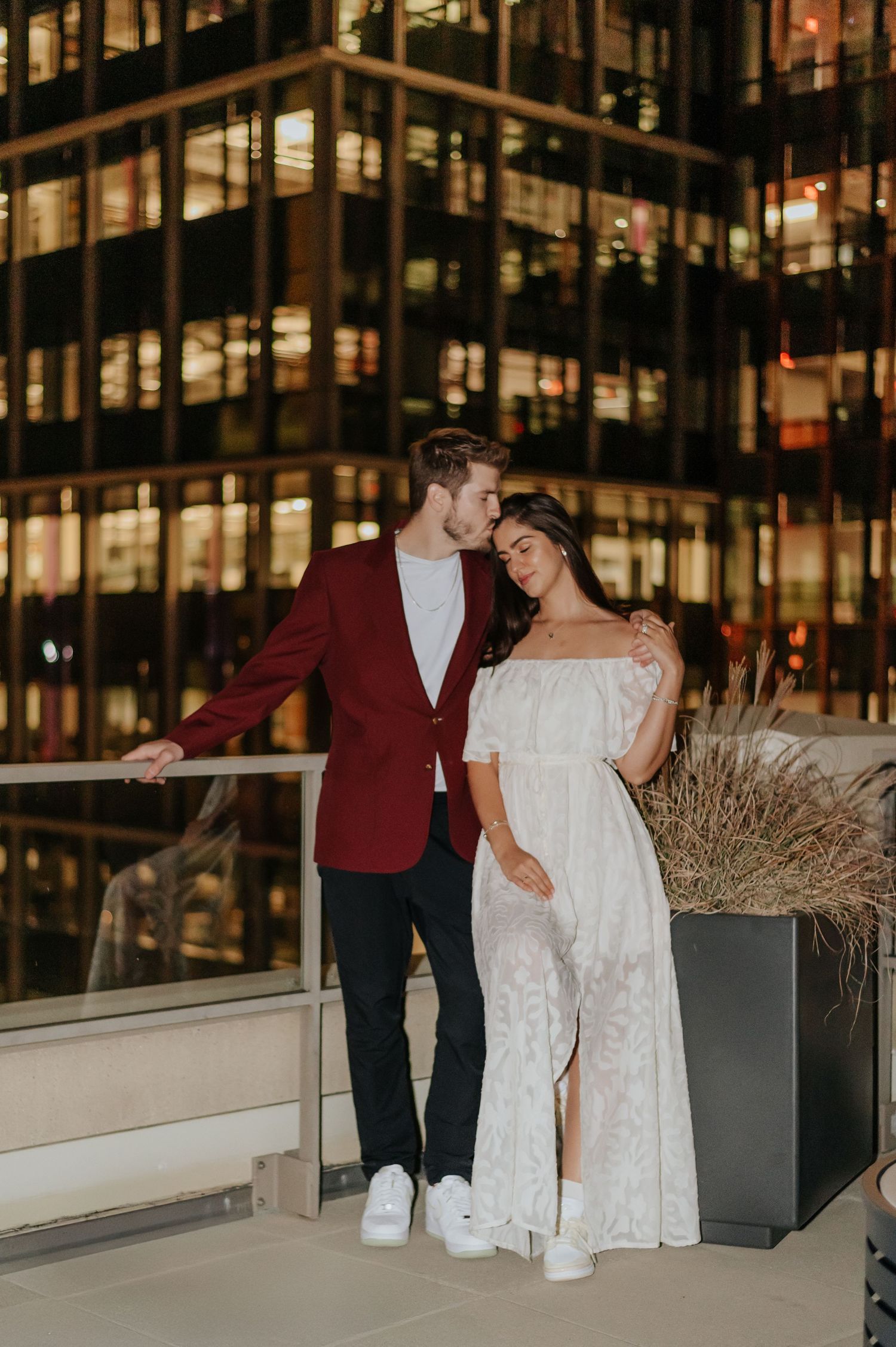 Couple in formal attire poses on rooftop terrace at night with illuminated office building in background.