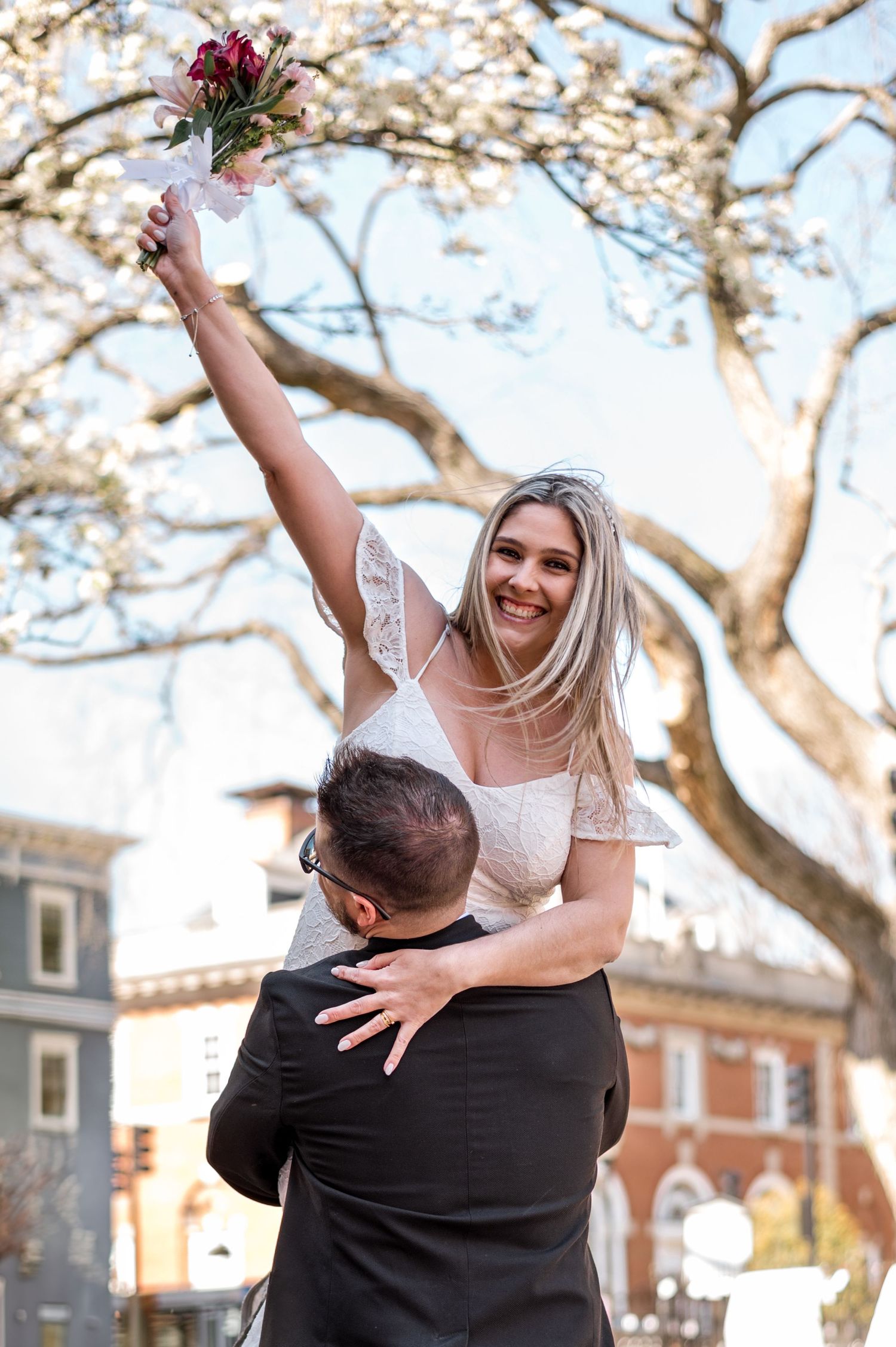 Celebratory lift pose with bride holding bouquet against blooming trees and historic architecture.