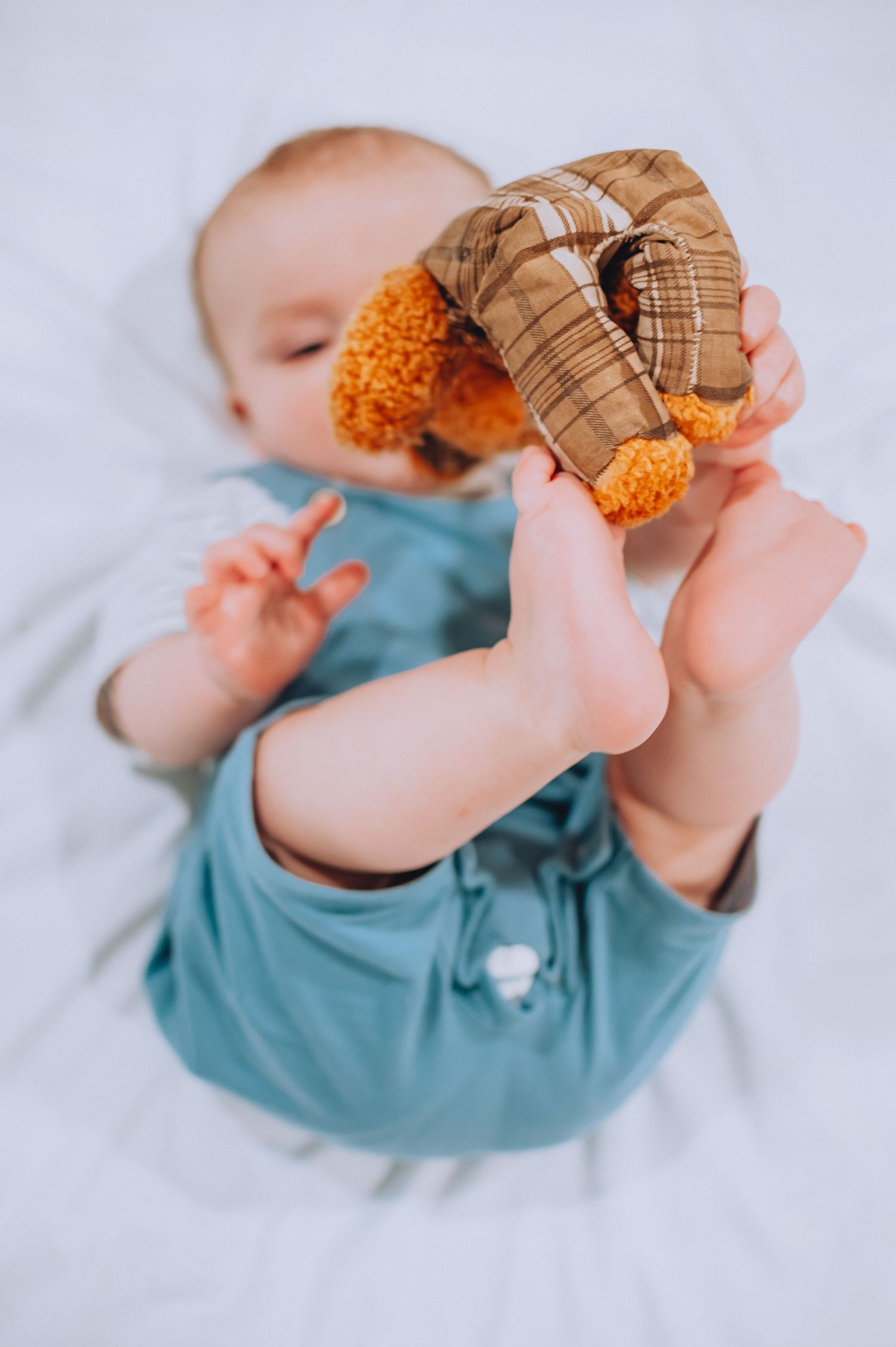 Infant in teal outfit playing with plush toy while lying on white blanket.