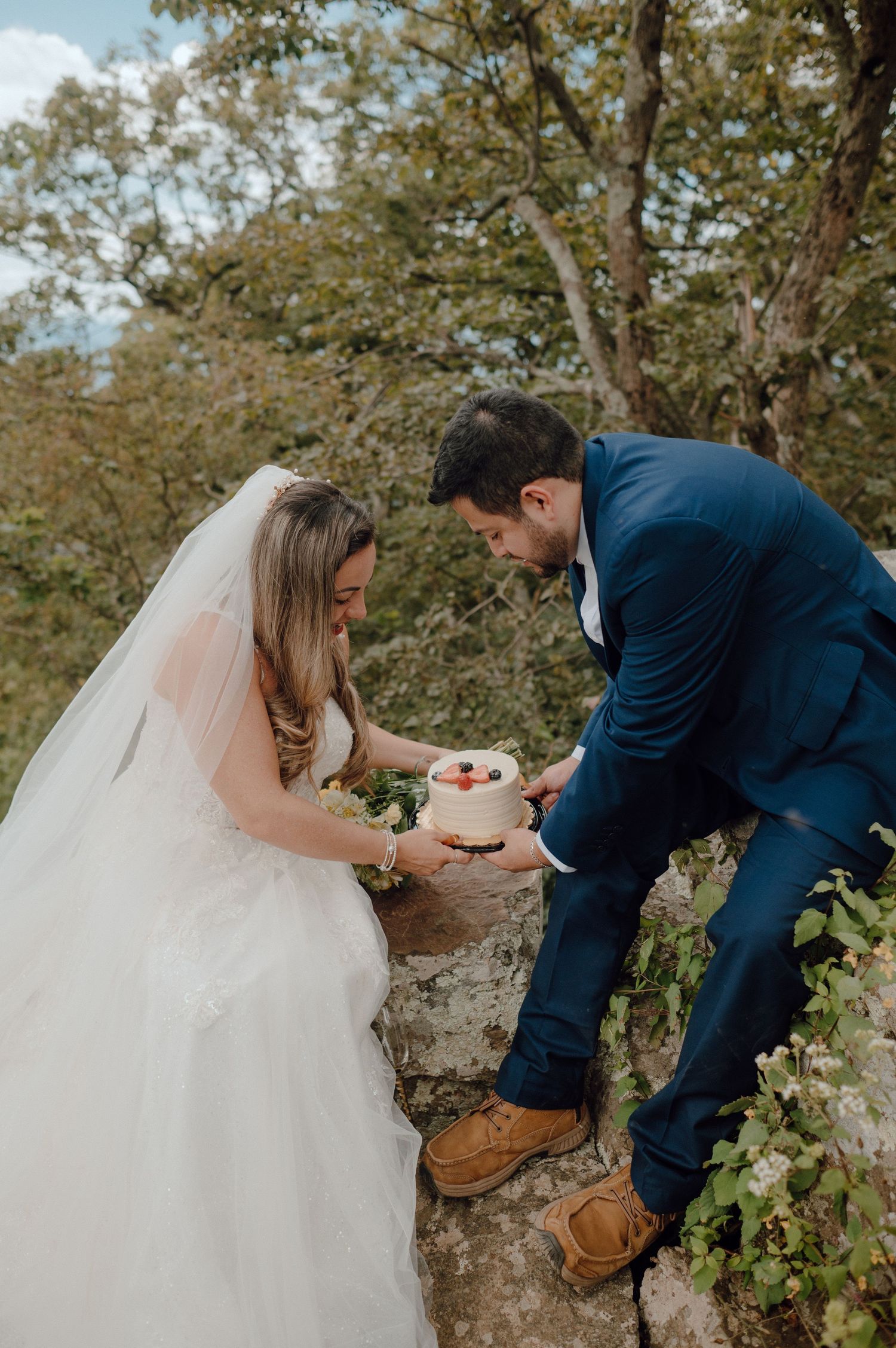Wedding couple shares an intimate moment on a rocky mountain ledge while handling something together.