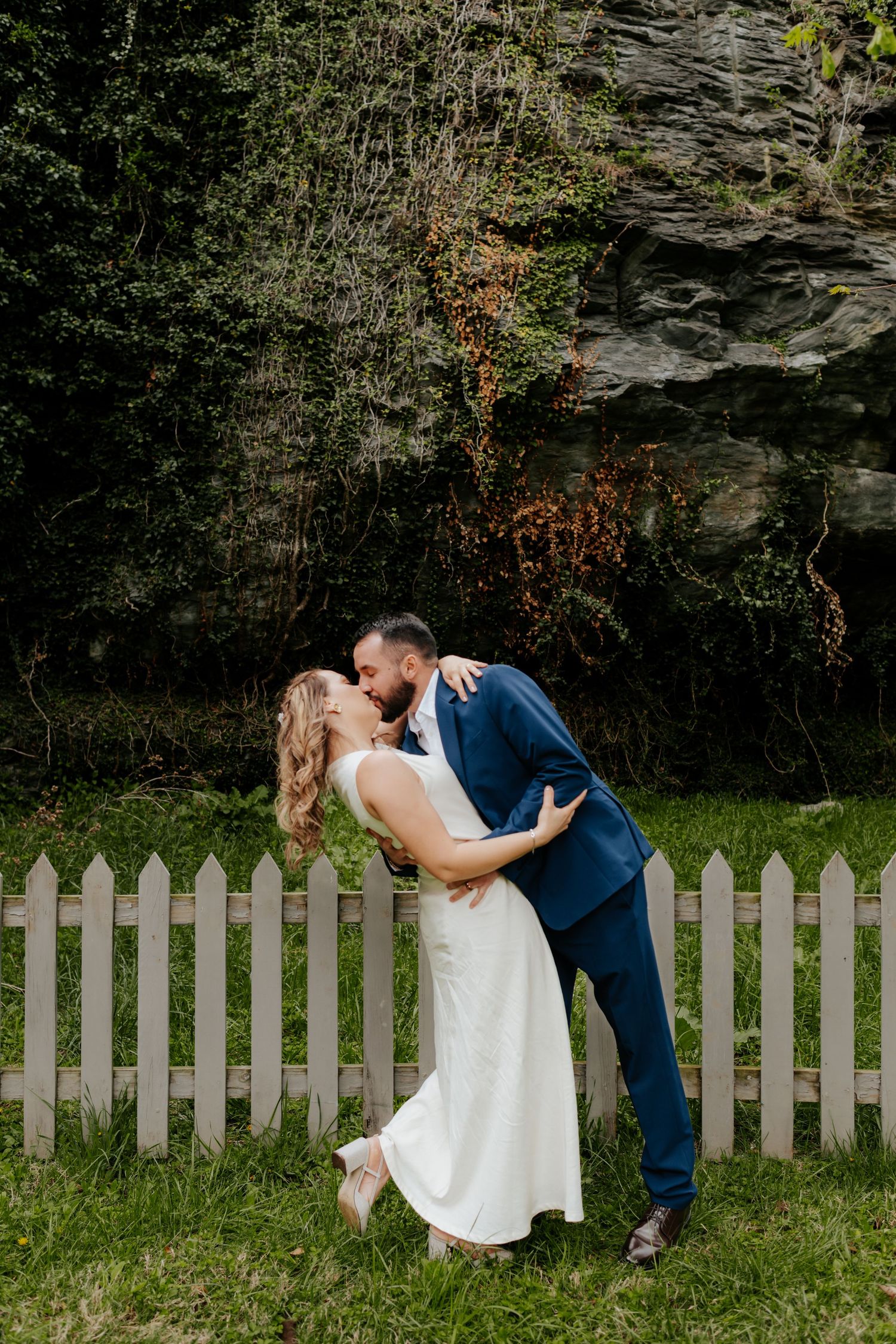 A romantic couple in wedding attire share an intimate dance pose against a stone wall and white picket fence backdrop.