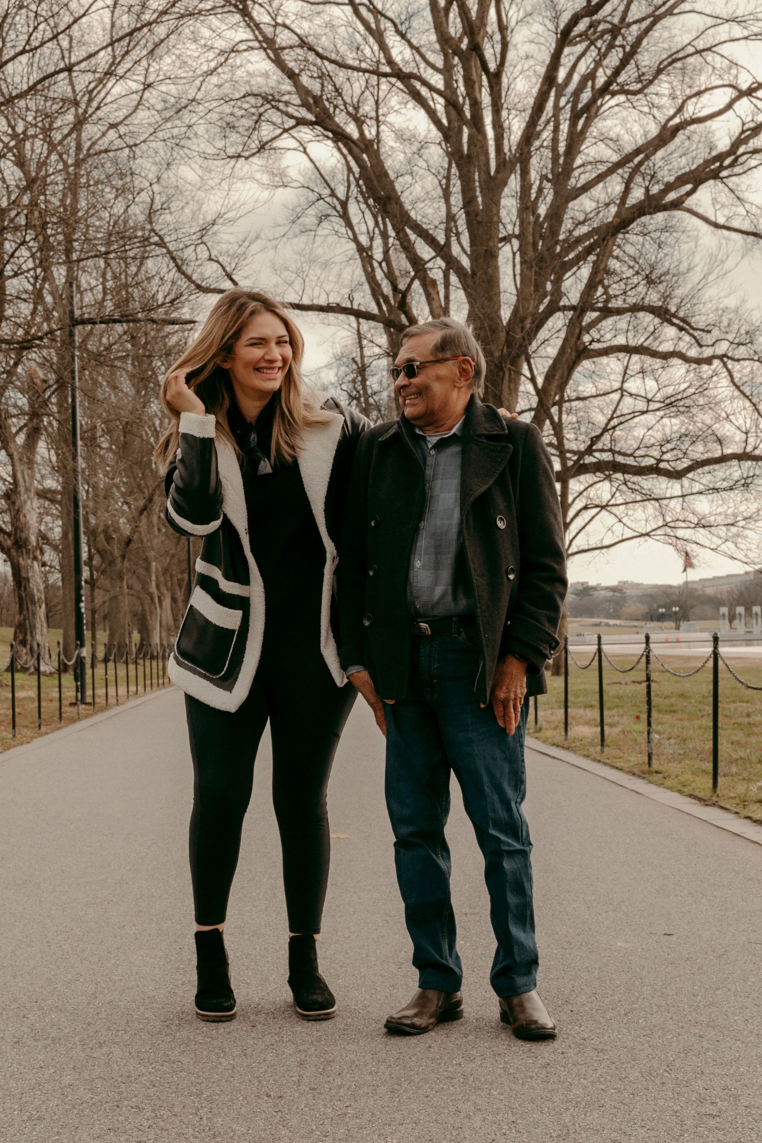 Two people in winter coats walking together along a tree-lined path.