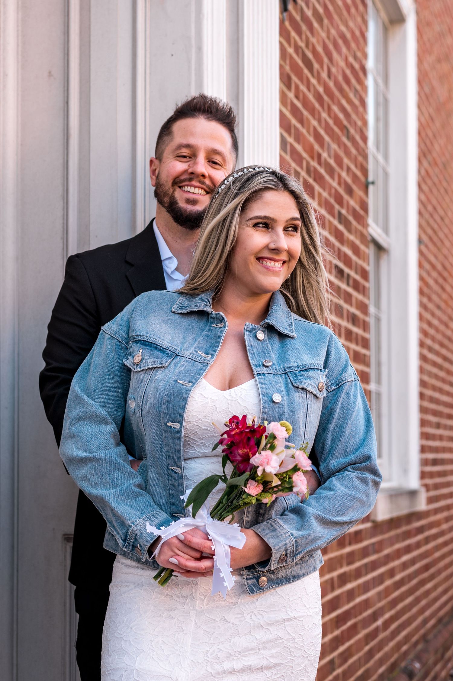 A couple poses for a portrait against a white wall, bride wearing a denim jacket over her wedding dress.