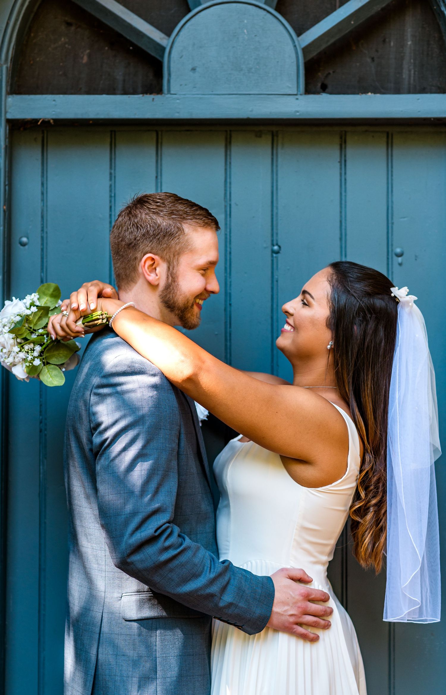 A romantic wedding portrait showing a couple embracing against a blue door with the bride holding a floral bouquet.