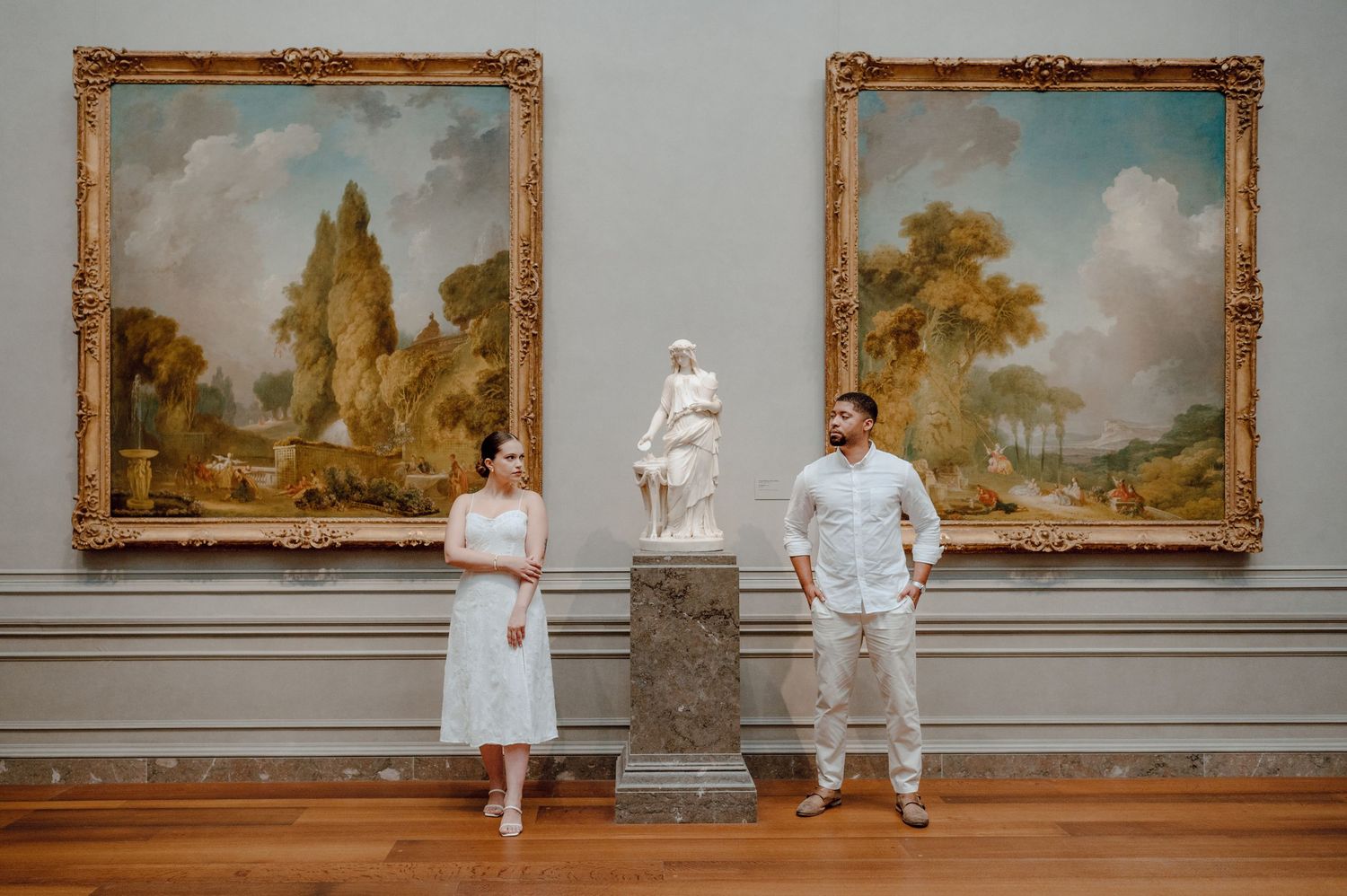 Two people stand in an art gallery between ornate gold-framed landscape paintings and a white marble statue on a pedestal.