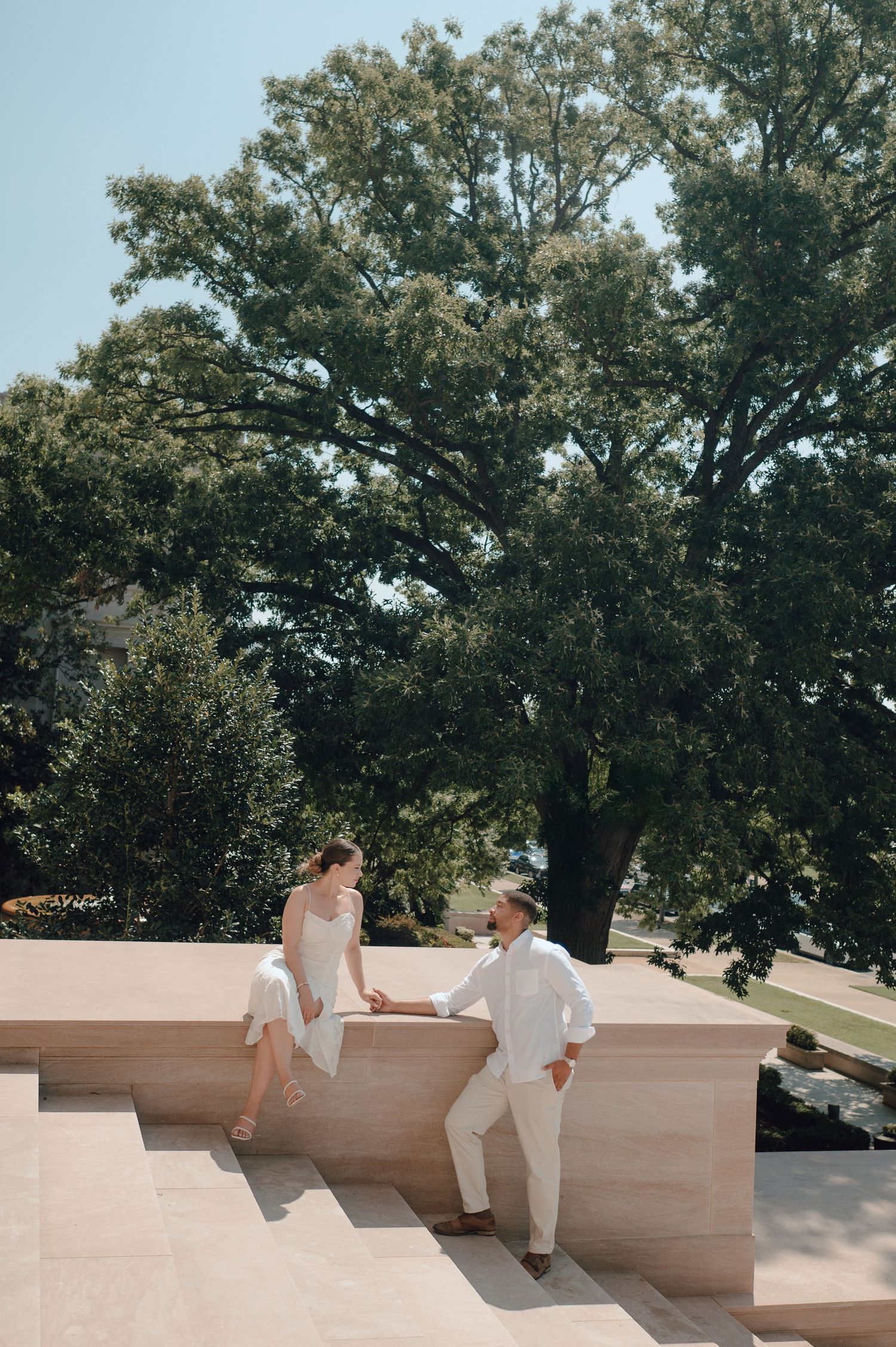 A couple stands on stone steps outdoors with lush green trees providing a natural backdrop on a sunny day.