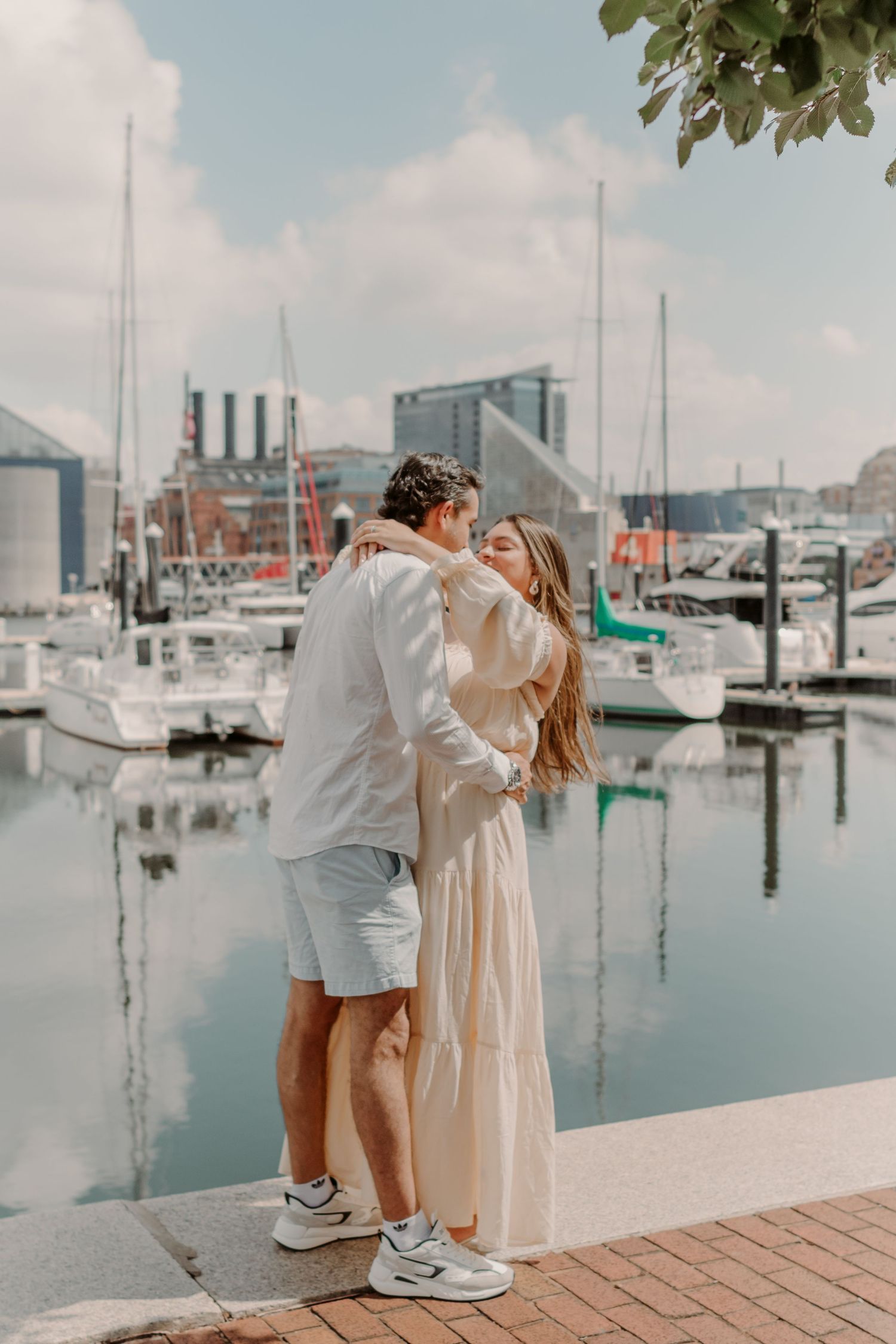 A romantic couple shares a tender moment on a brick pathway by a marina filled with boats on a sunny day.