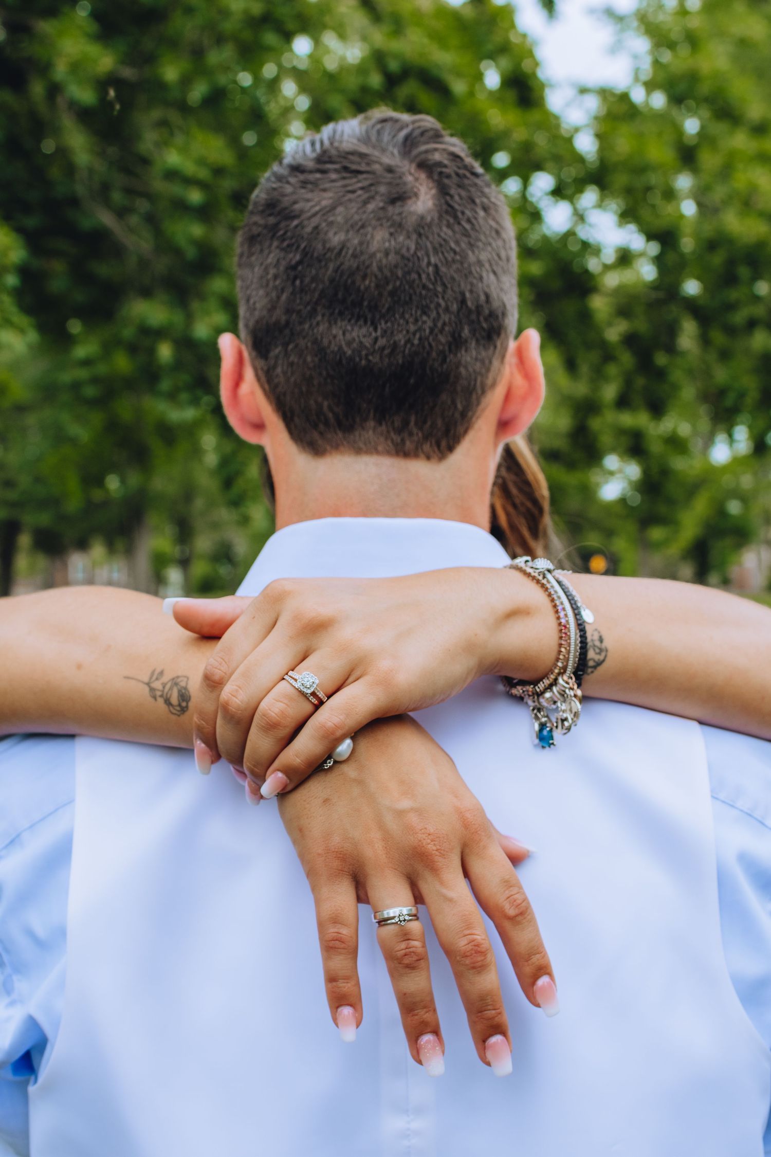 Close-up of hands with jewelry and tattoo embracing someone wearing a light blue shirt outdoors with green foliage.