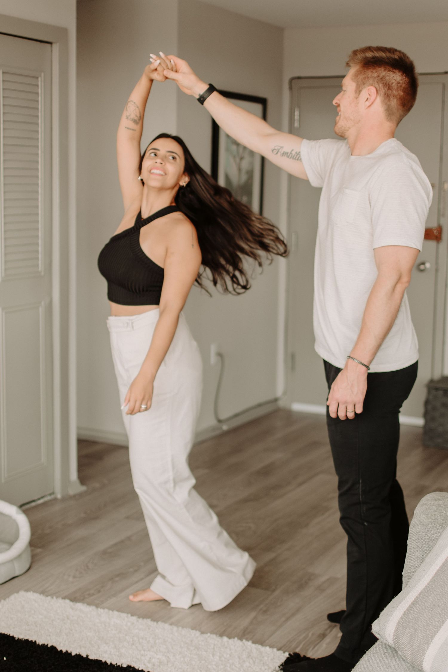 A couple dances together in a bright living room, with the woman twirling in flowy white pants and a black top.