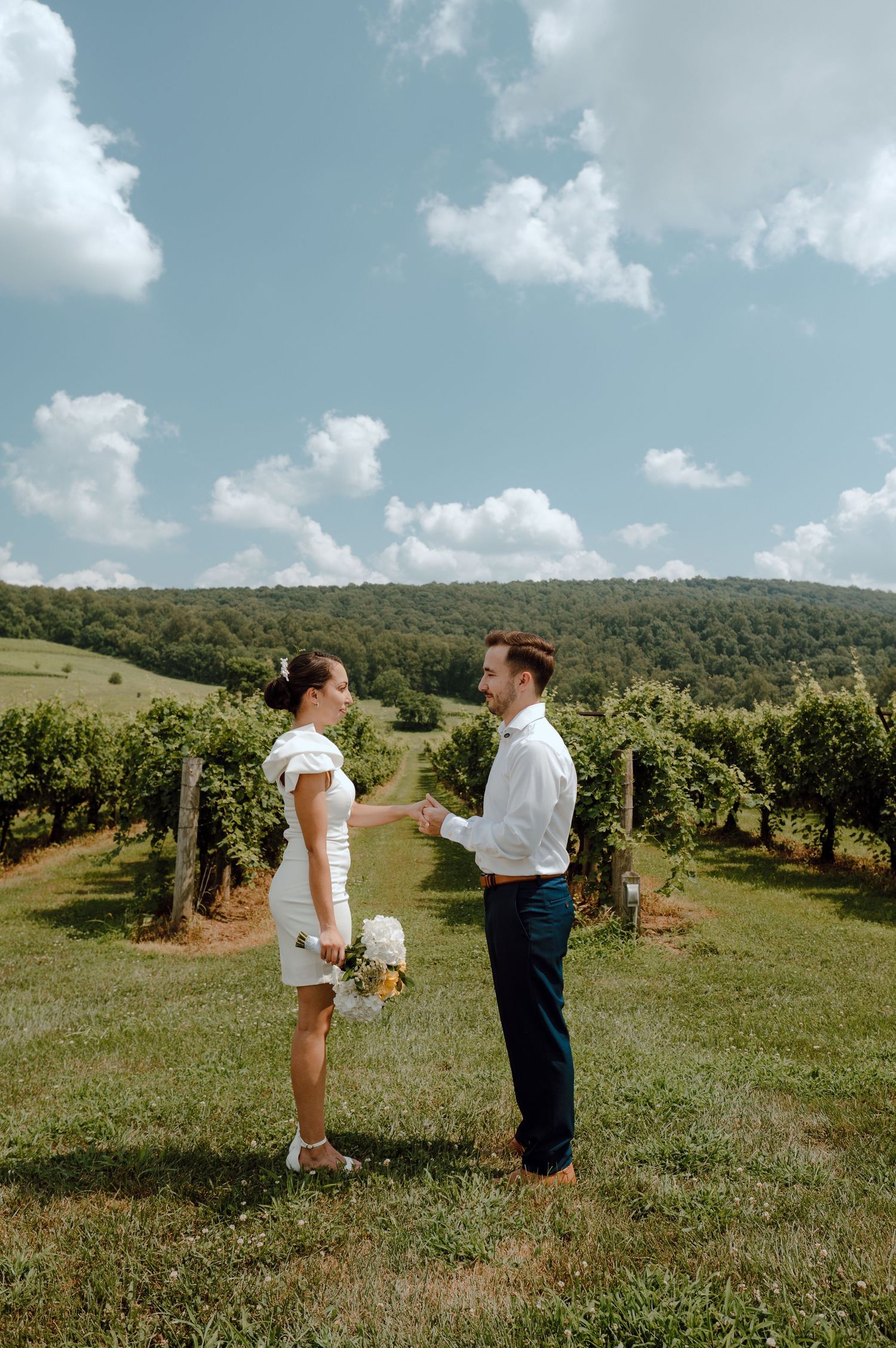 Two people stand together in an outdoor vineyard setting on a sunny day with rolling hills in the background.
