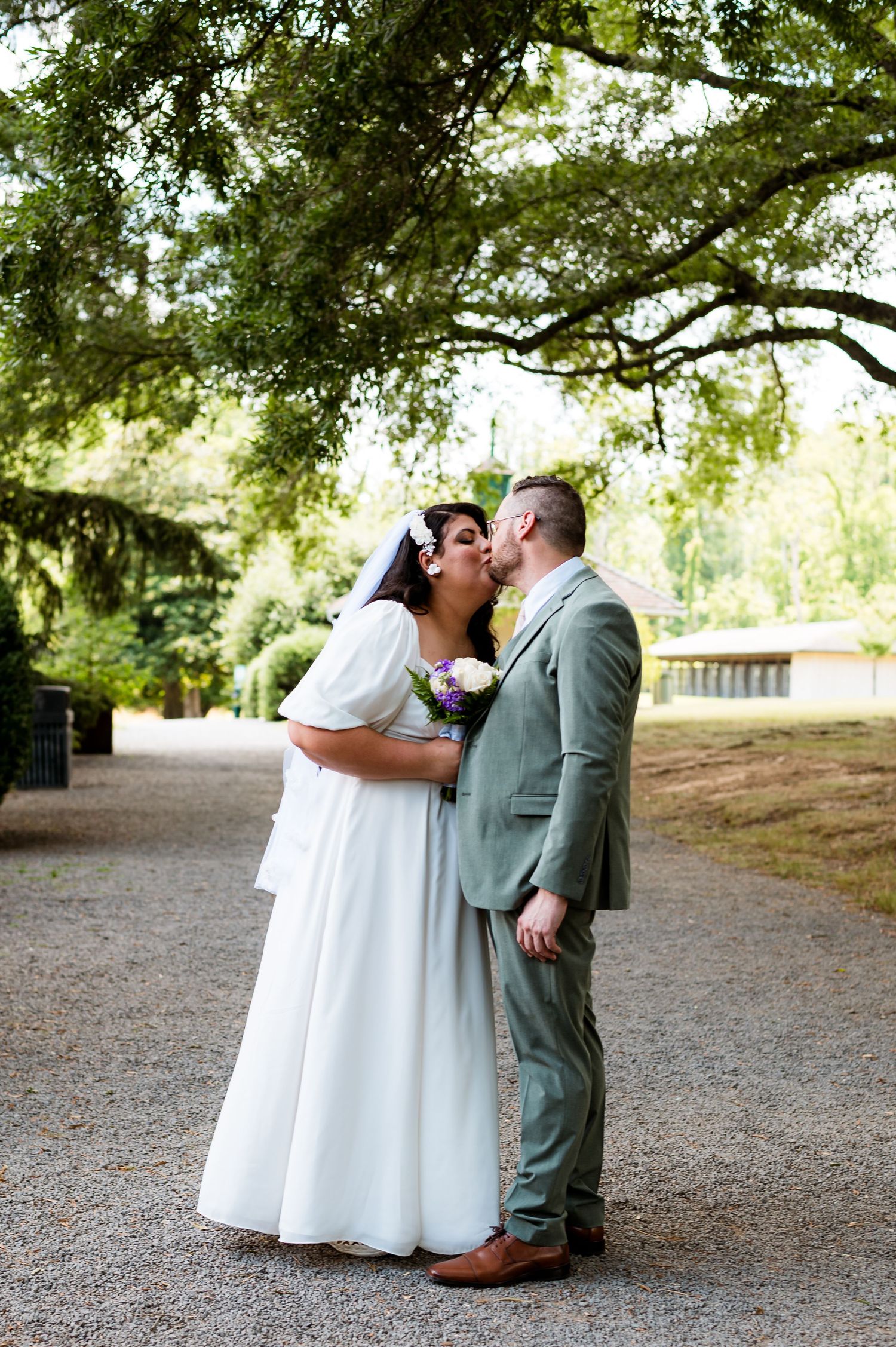 A wedding couple shares an intimate moment under green trees on a gravel path in a garden setting.