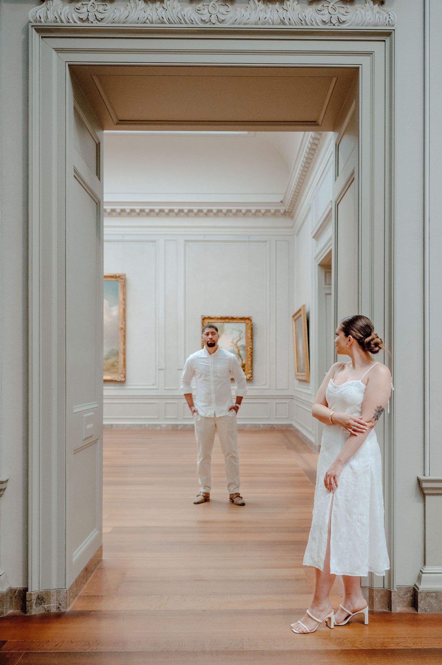 Couple in white attire poses elegantly in an art gallery hallway with ornate moldings and wooden floors.