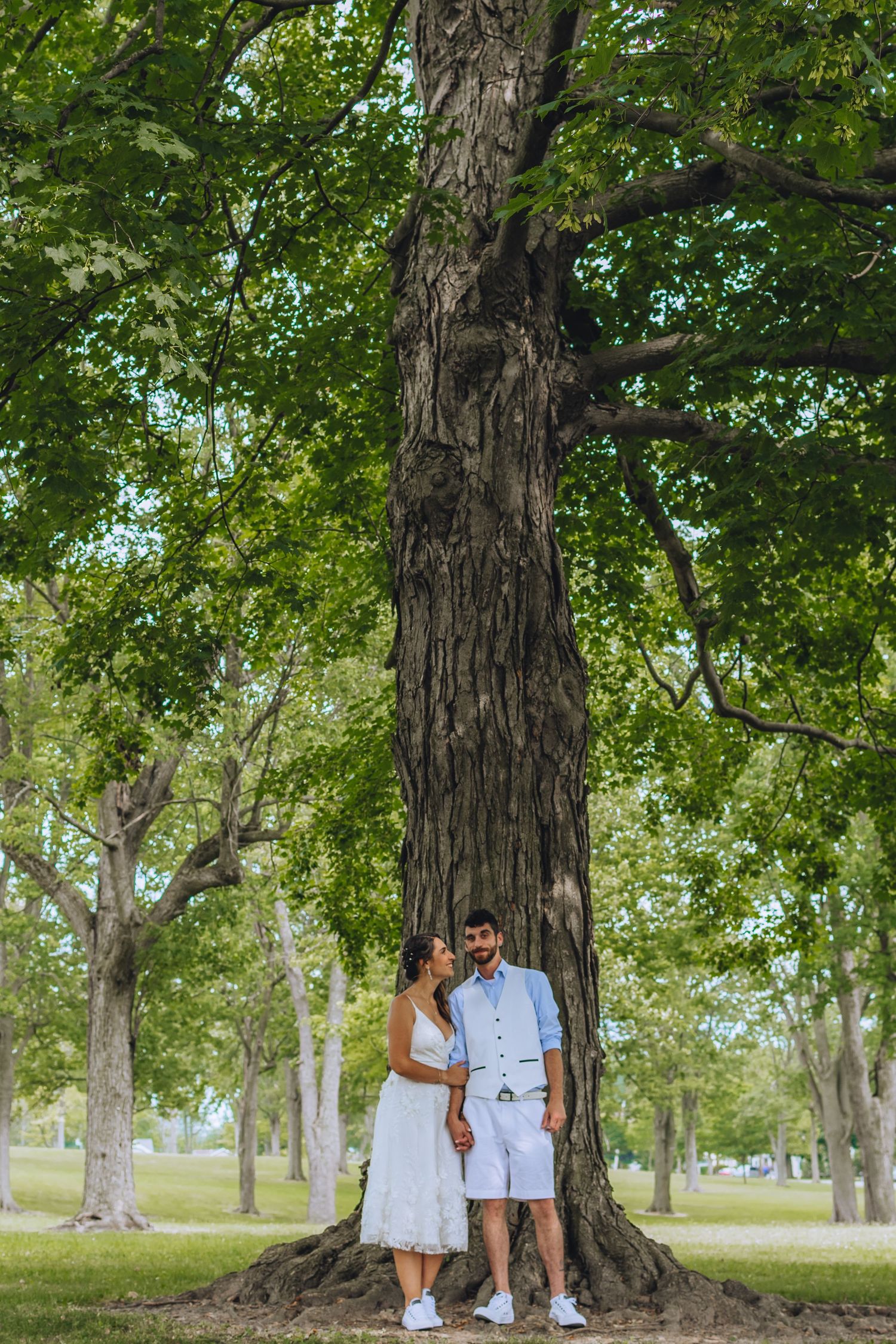 A couple stands together under large trees in a park during a sunny day wearing coordinated white and blue outfits.