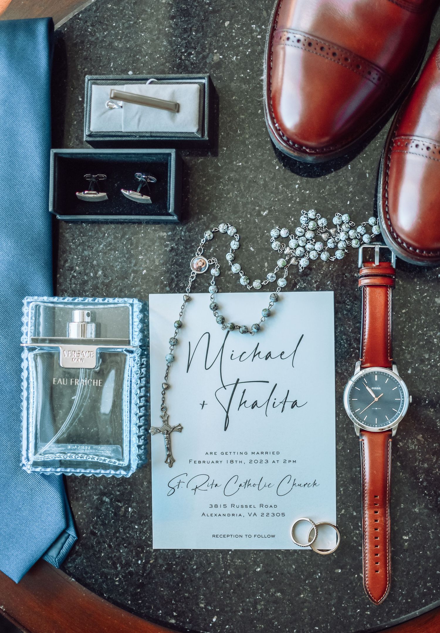 Wedding details flatlay with brown leather shoes, cologne, watch, jewelry, and invitation card on dark background.