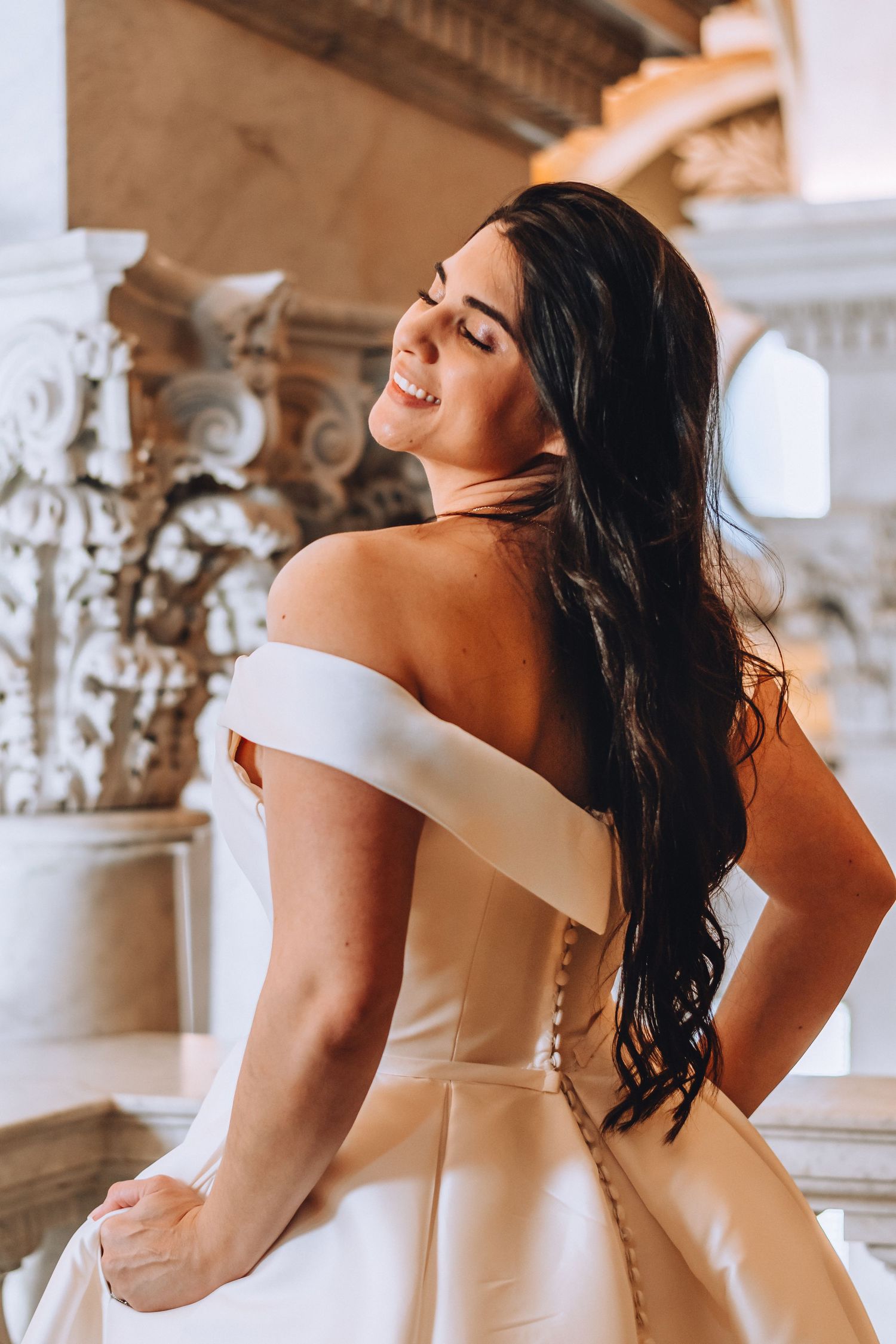 A bride in an off-shoulder white gown poses with her back turned, showing intricate dress details.
