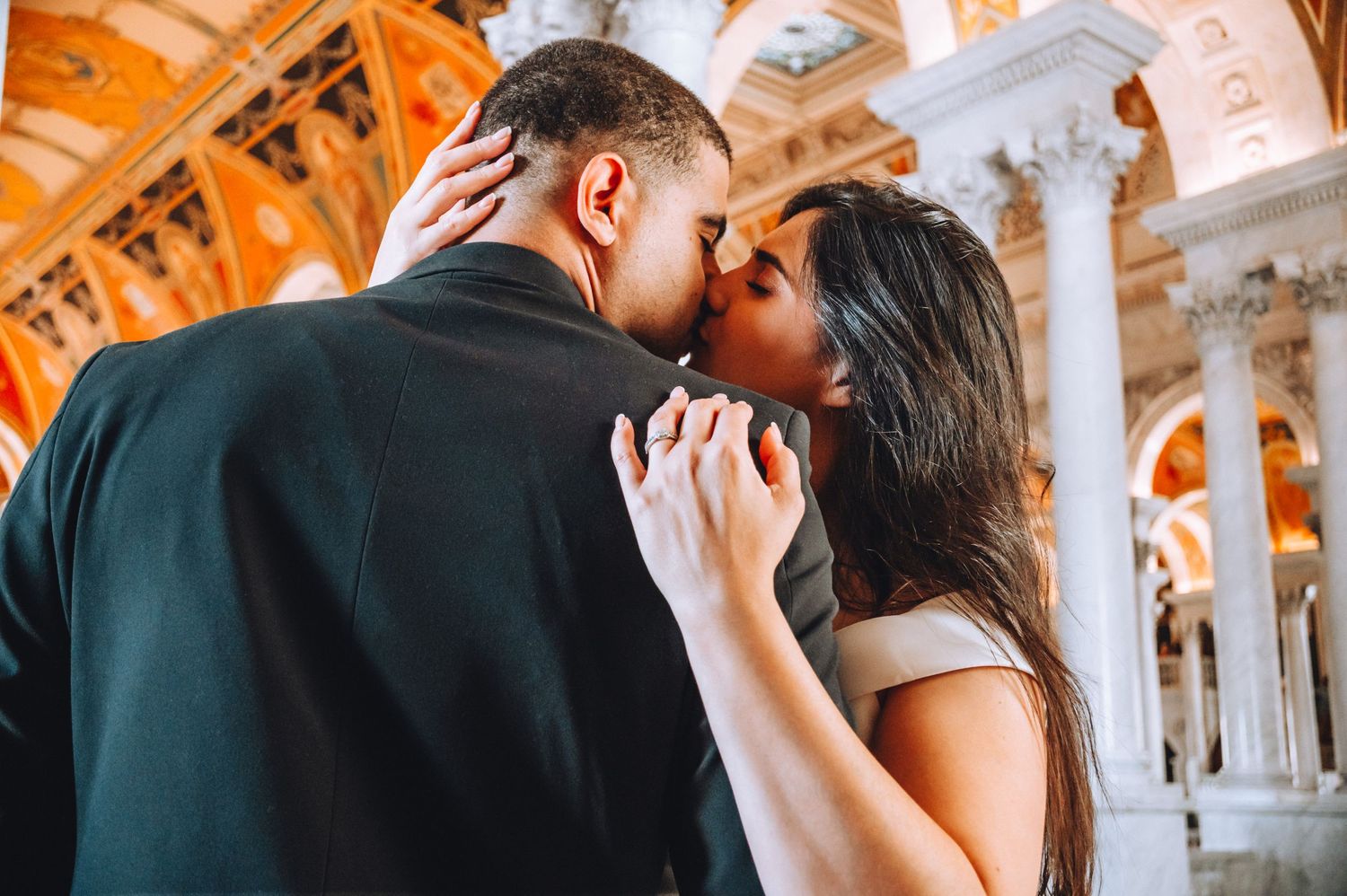 A close-up romantic moment between newlyweds with ornate orange-tinted architectural details in background.