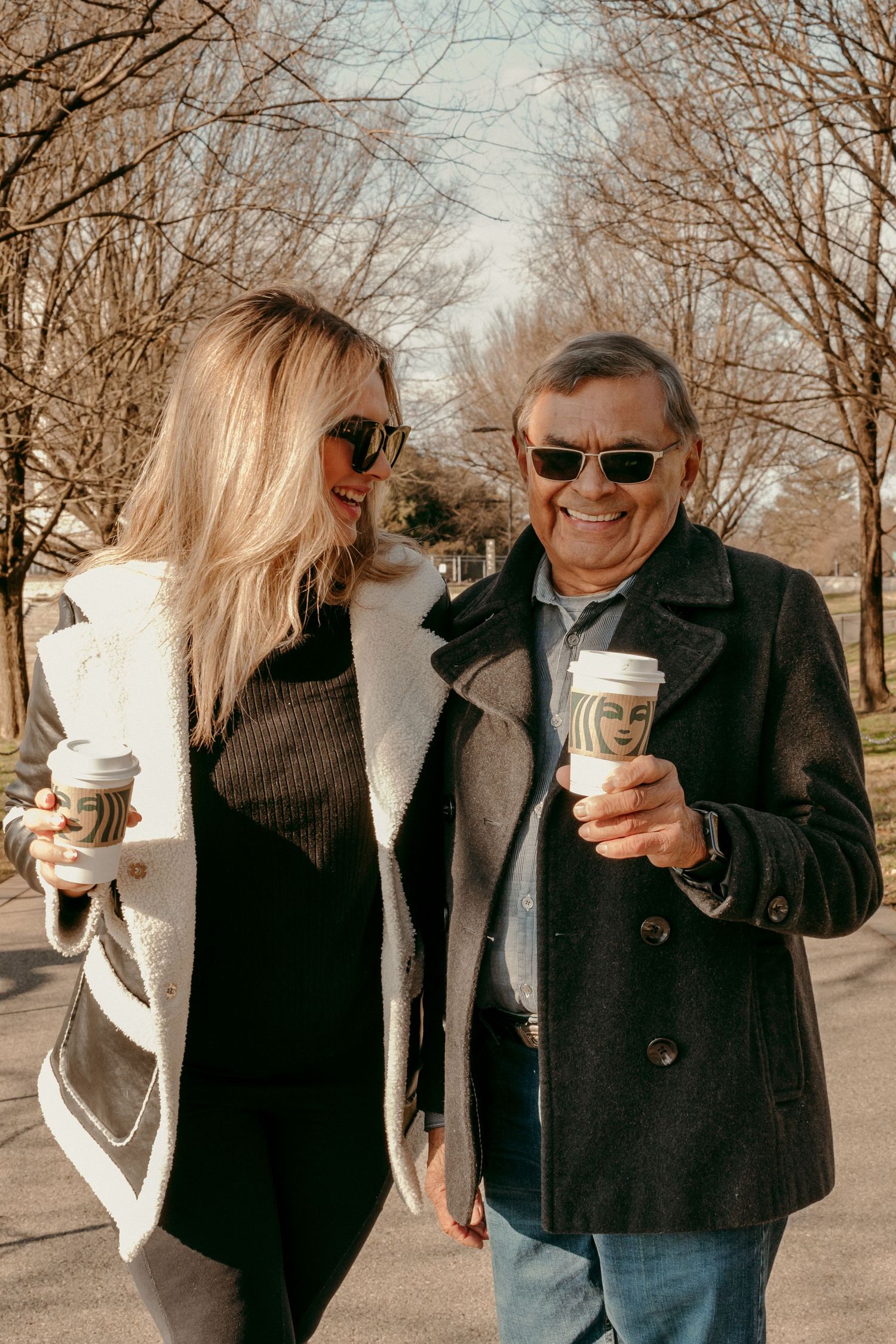 Two people in winter coats holding coffee cups while walking outdoors on a sunny day.